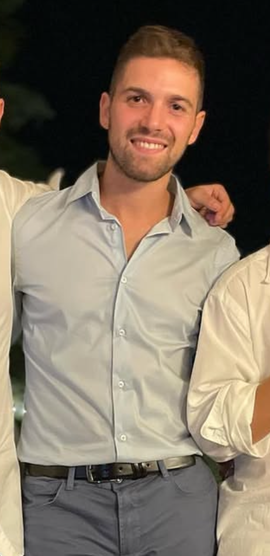 Un uomo sorridente seduto in un'auto, indossando una maglietta blu con scritte, con la mano sollevata in un gesto amichevole.