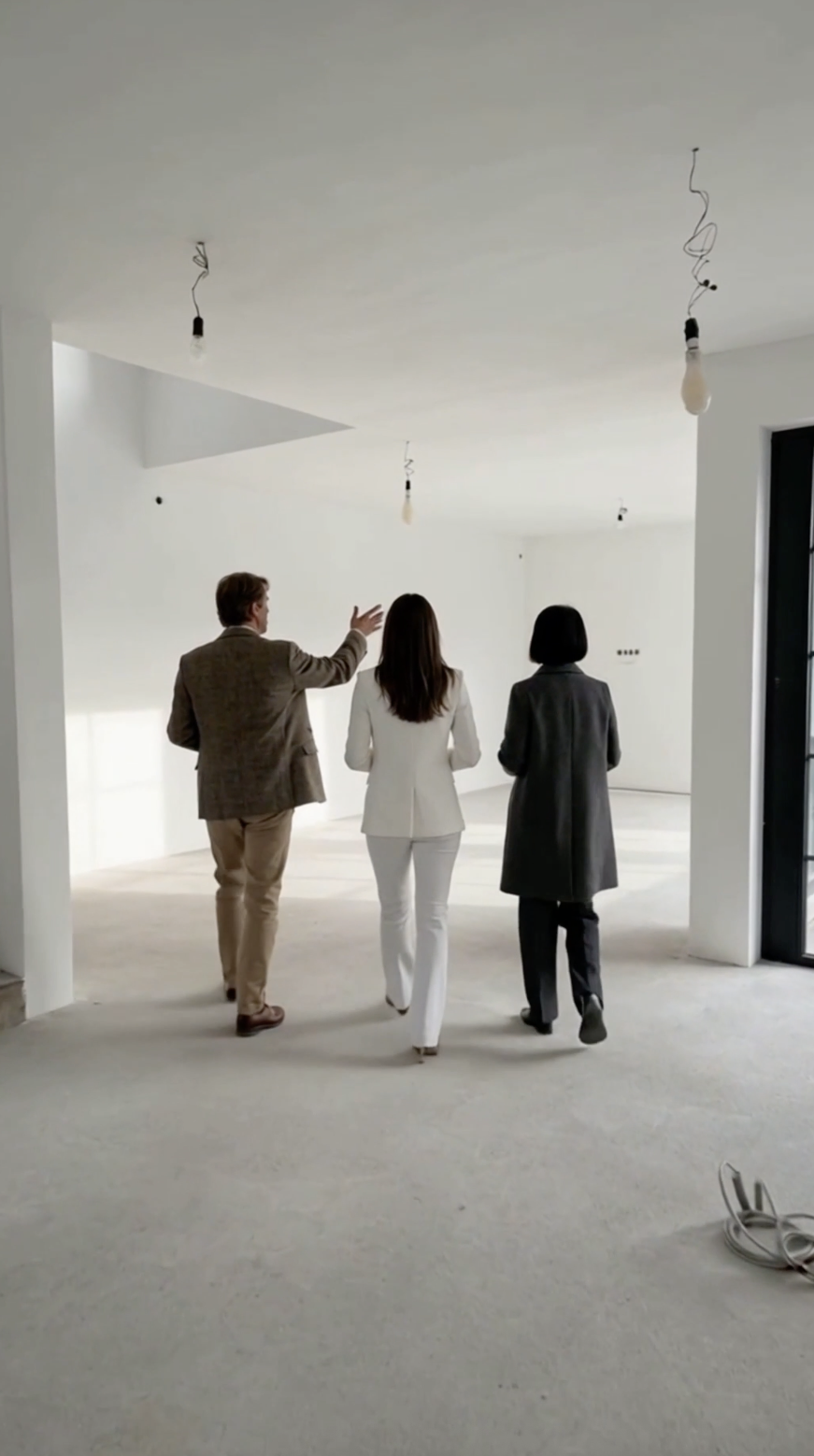 Tre persone che camminano in un ambiente vuoto e moderno, probabilmente un appartamento o un ufficio, con pareti bianche e luci pendenti non ancora installate.