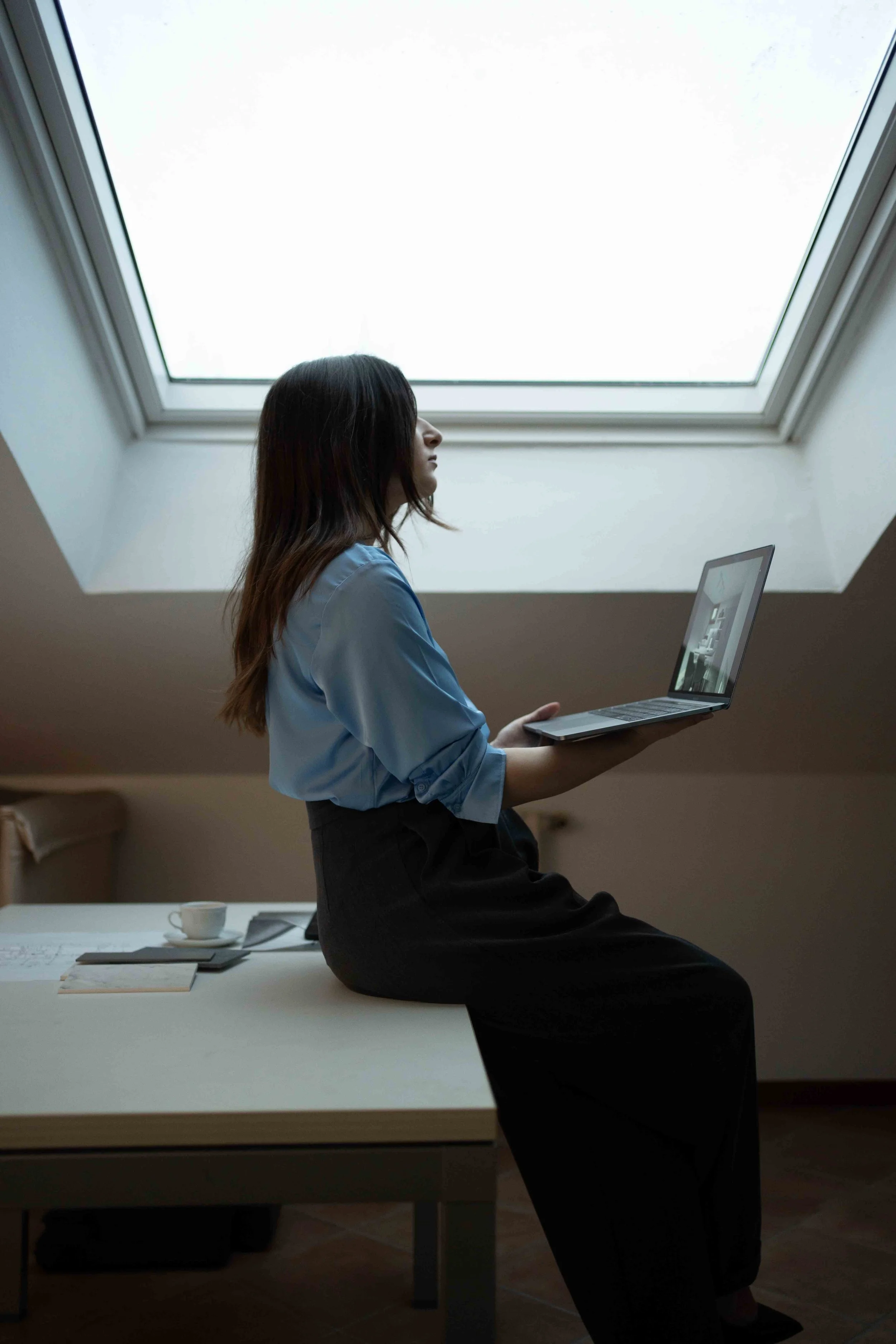 Donna seduta su un tavolo con un laptop in mano sotto una grande finestra da soffitto