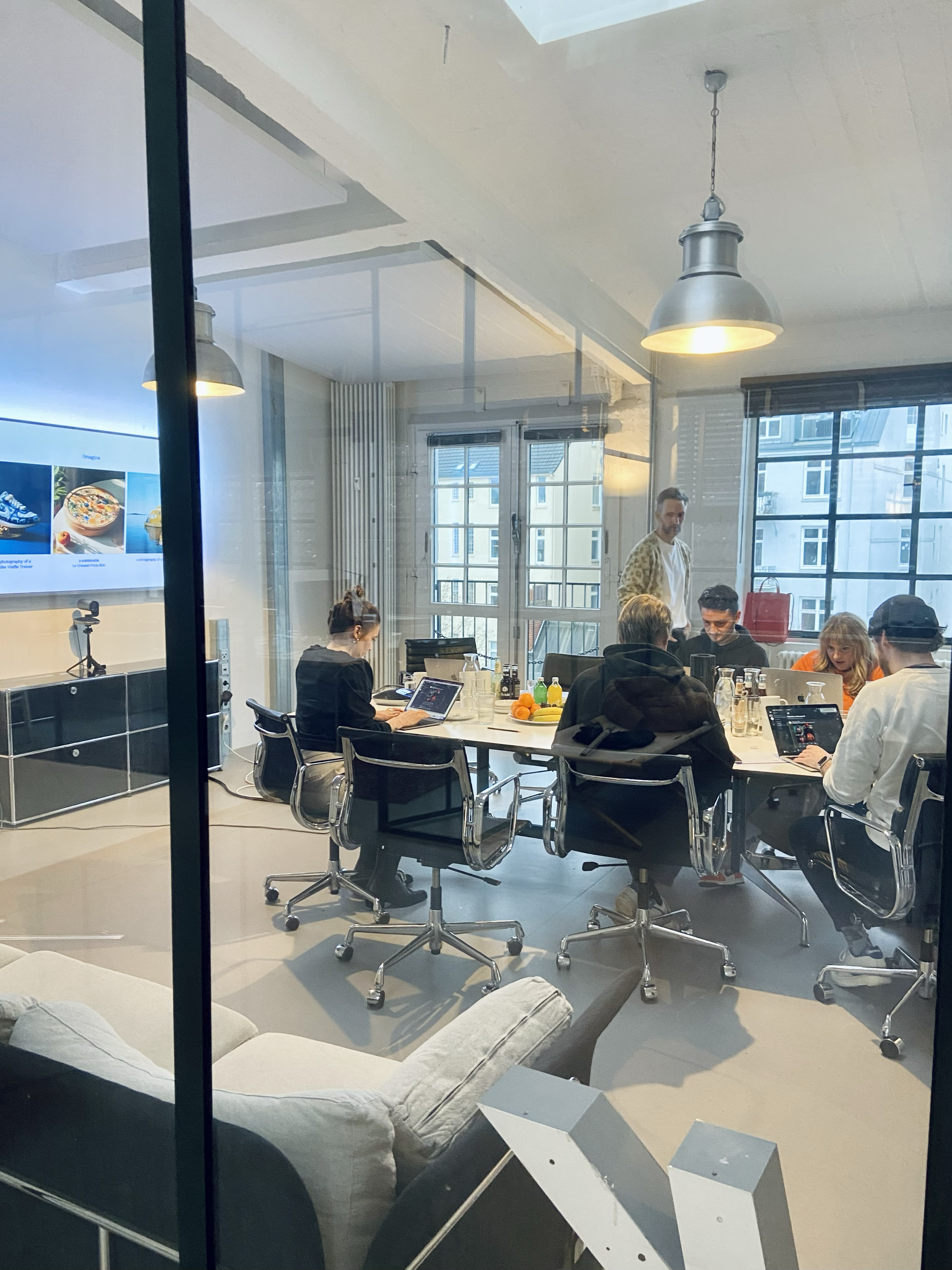 Blick durch eine Glasscheibe auf ein modernes Büro mit mehreren Menschen, die am Tisch arbeiten, und einem Mann, der steht. Es gibt große Fenster, eine Deckenleuchte, und einen Bildschirm an der Wand im Hintergrund.