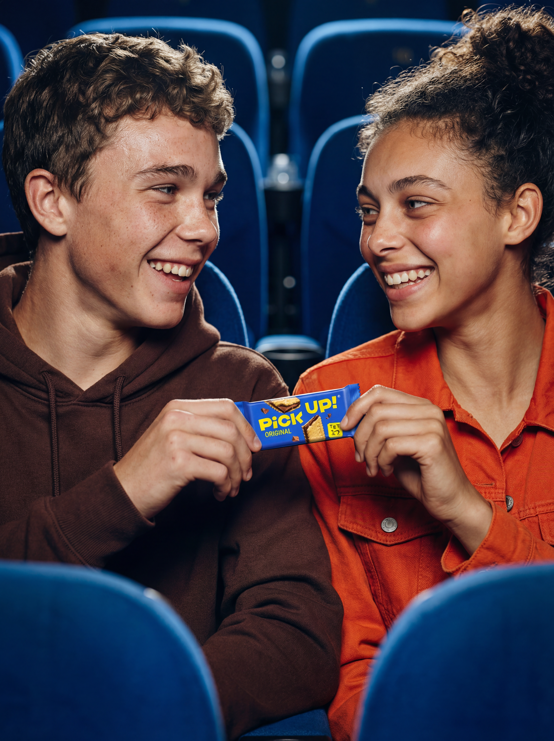 Zwei junge Menschen, ein Mann und eine Frau, sitzen in einem Kinosaal und lachen miteinander, während sie eine Schokoriegel teilen.