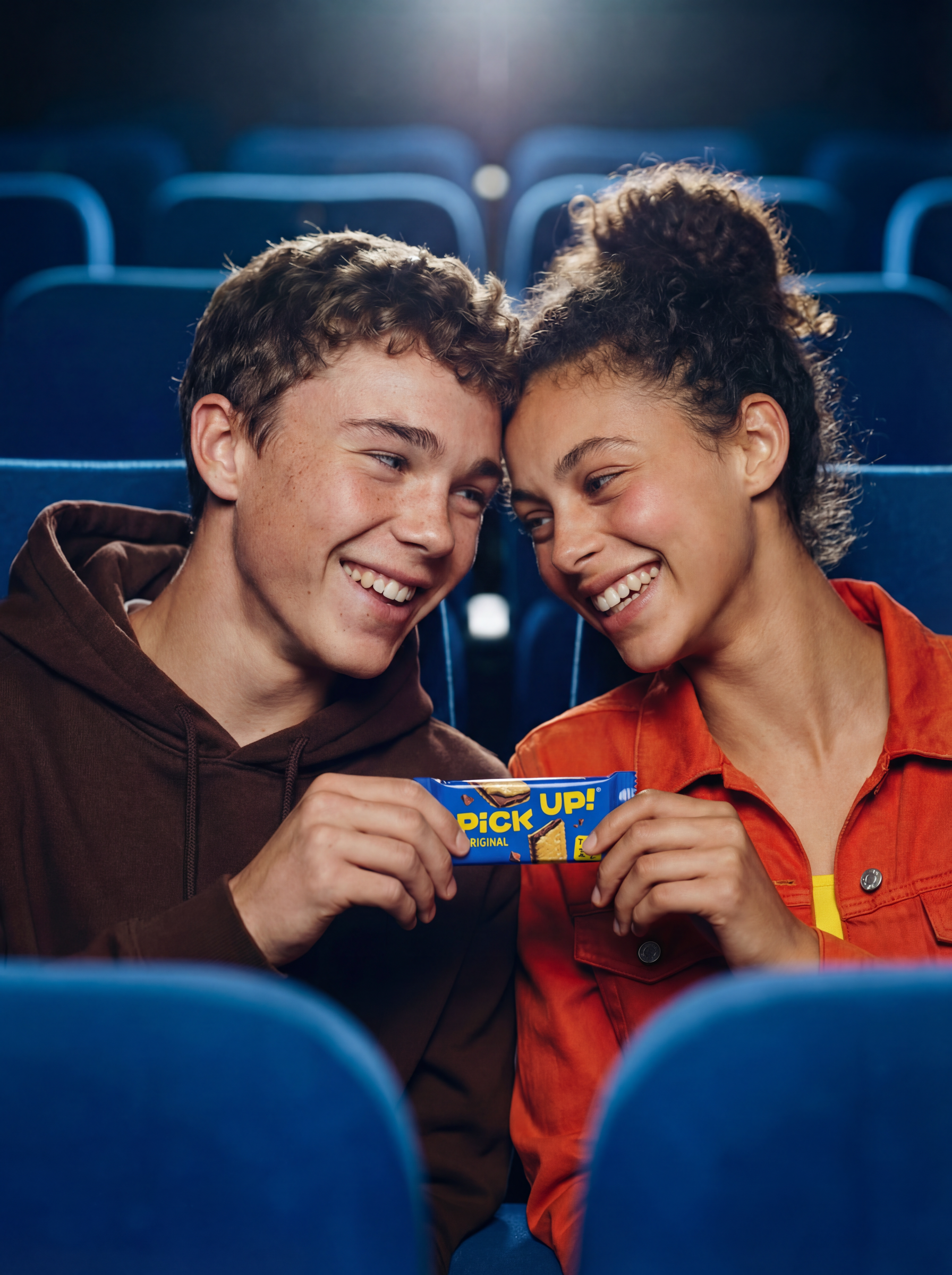 Zwei junge Erwachsene sitzen in einem Kinosaal, lachen und teilen sich einen Snack.