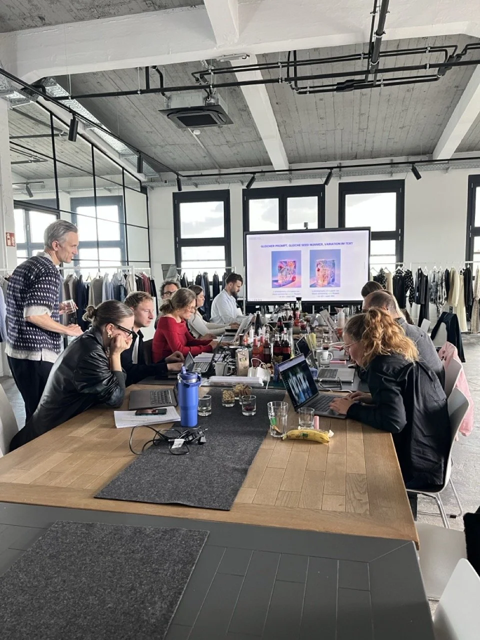 Eine Gruppe von Menschen arbeitet in einem modernen, lichtdurchfluteten Konferenzraum mit großen Fenstern, einem großen Holztisch, Laptops, Getränken und einem großen Bildschirm, der Präsentationsfolien zeigt.