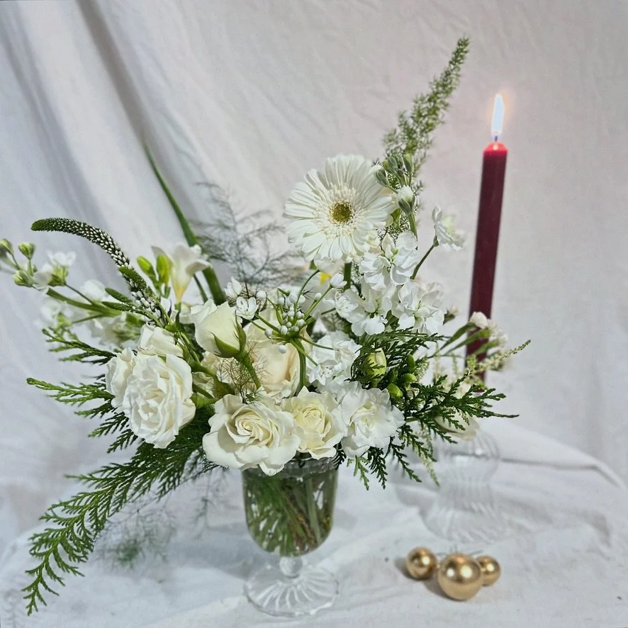 Timeless holiday floral arrangement with red ranunculus, white roses, irises, and lush greenery, available in classic red and white or elegant all-white designs, styled in a glass vase.