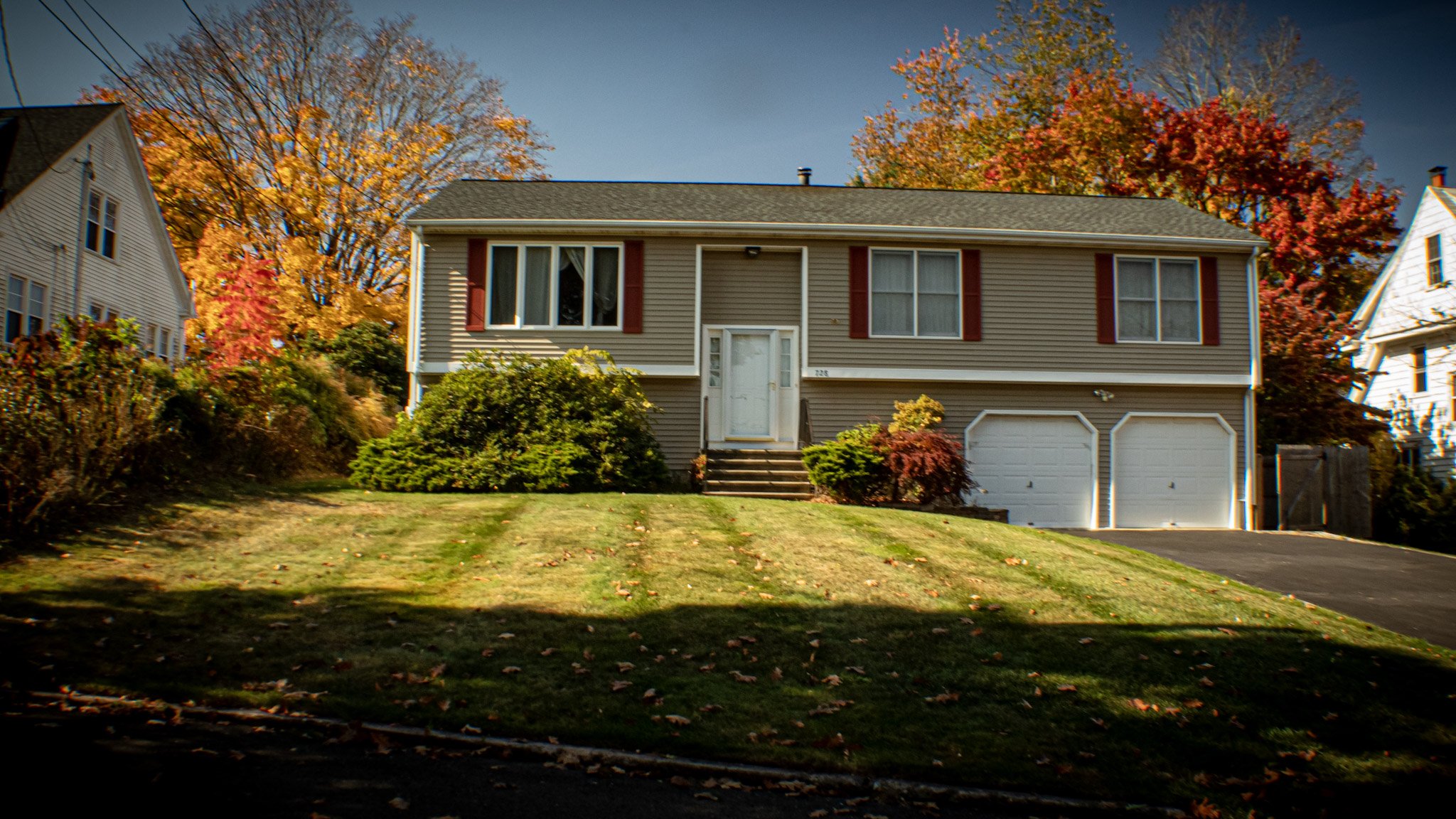 A beige two-story house with a front lawn and trees with fall foliage in the background. The house has a front door accessed by stairs and a two-car garage.