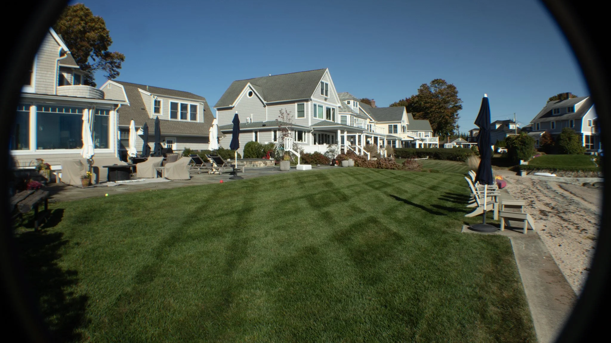 View of a backyard with a well-maintained lawn, patio chairs under umbrellas, and a row of houses in the background on a clear day.