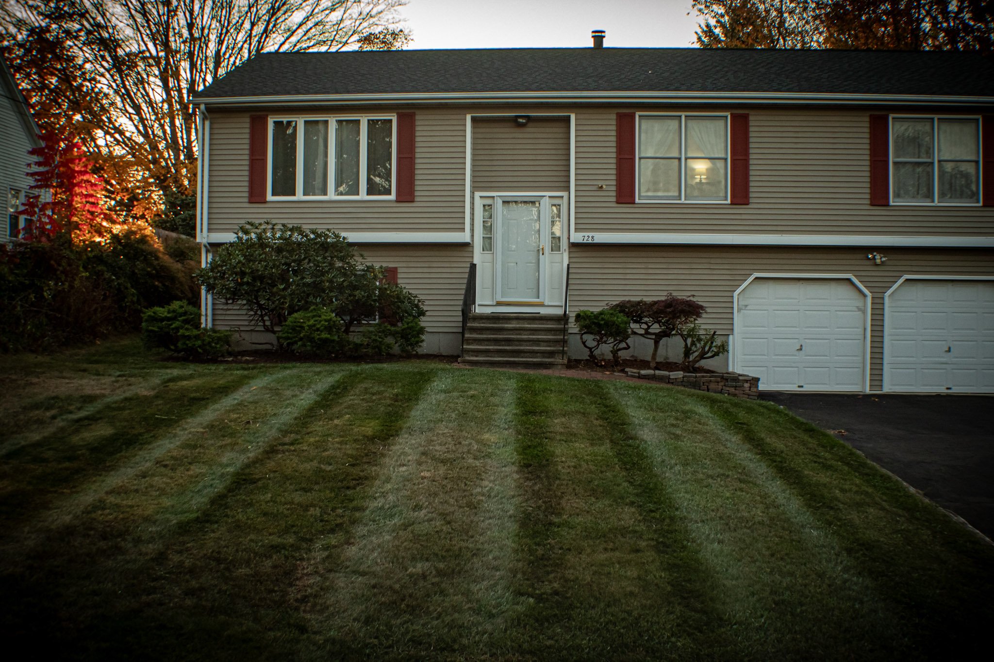 Front view of a two-story suburban house with beige siding, red shutters, a white front door, and a well-maintained lawn with striped grass. There are bushes and small trees in the yard, and a driveway with white garage doors on the right.