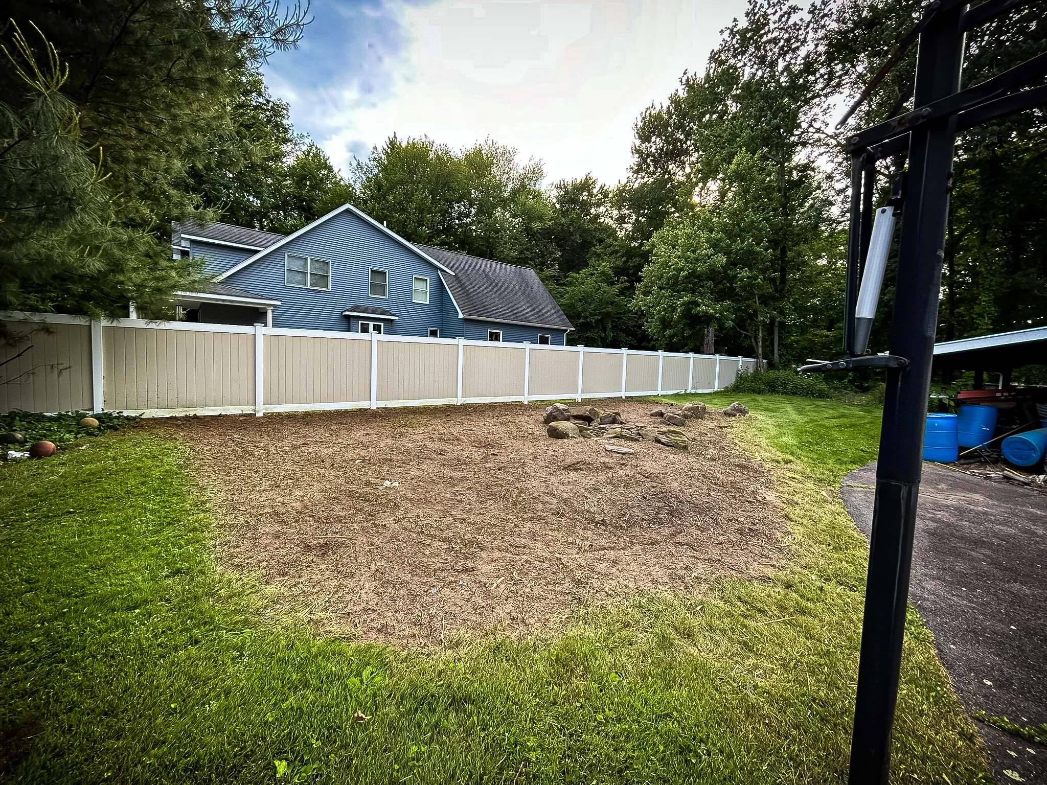 A backyard with partially seeded grass and a white vinyl fence with a blue house in the background. There are some rocks in the yard and a basketball hoop on the right side.