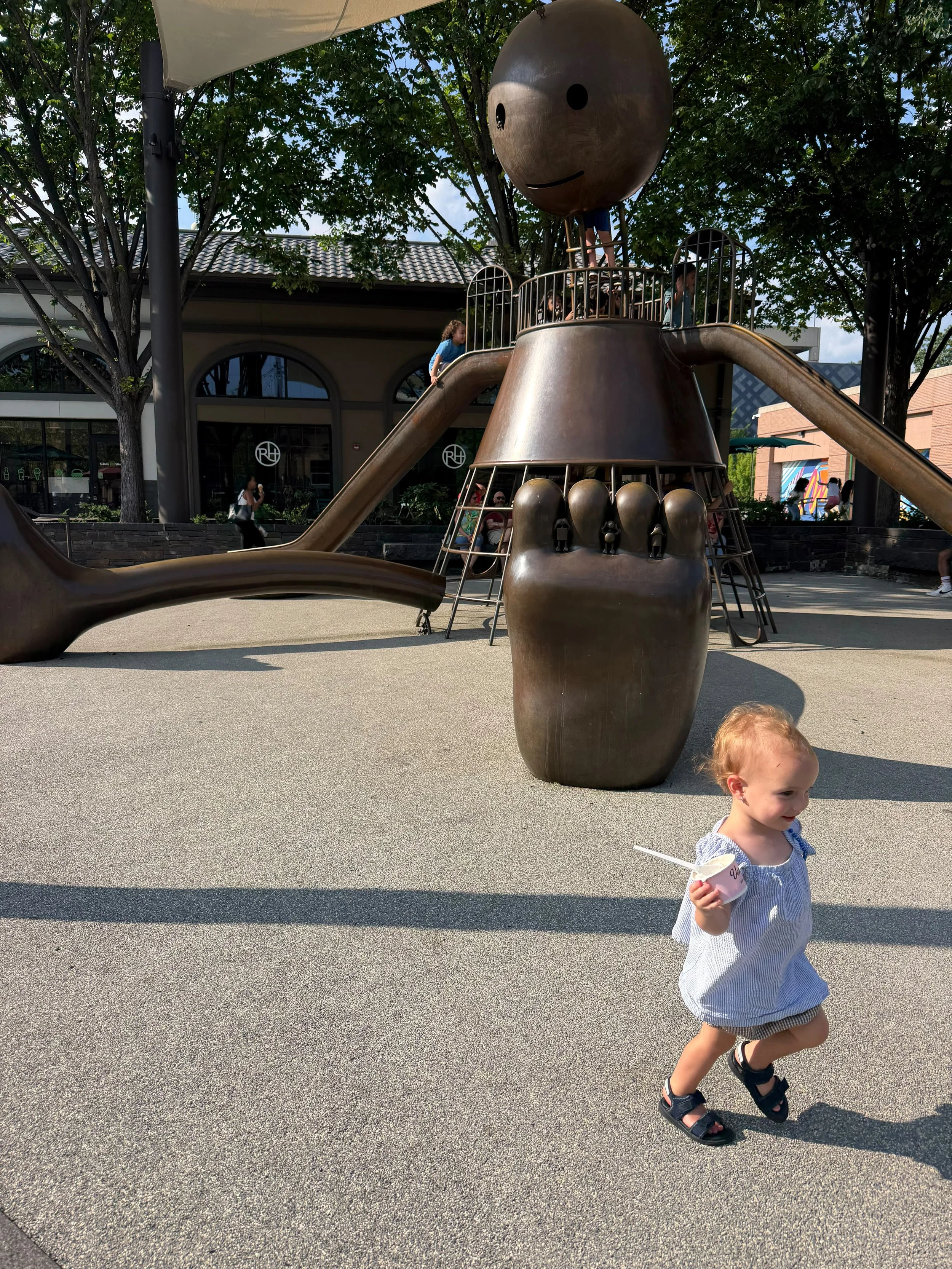 A toddler walking past a sculptural playground while holding ice cream, illustrating the limited play opportunities for younger children.