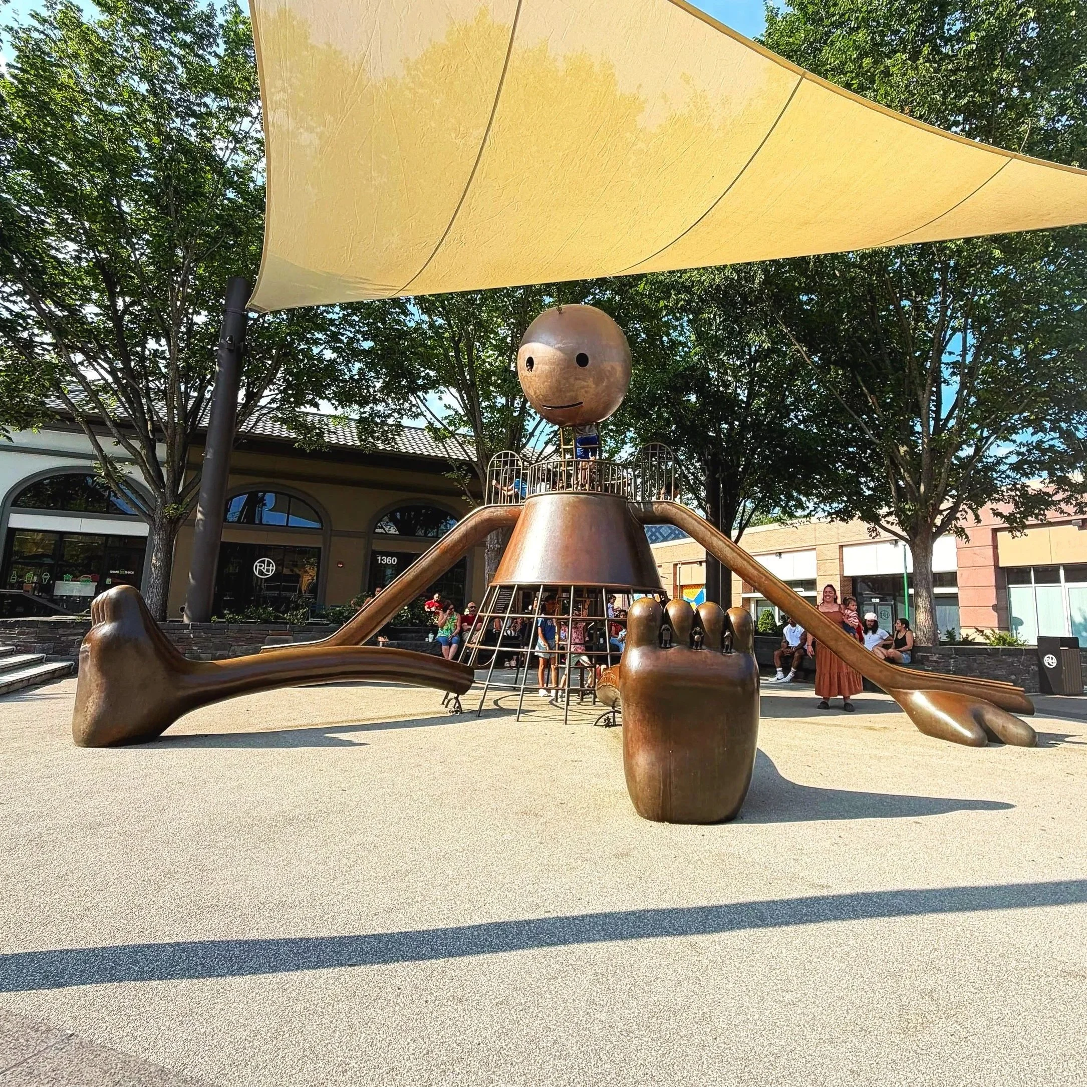 A sculptural bronze playground in a shopping center plaza featuring oversized slides, shade sails, and a giant seated figure.