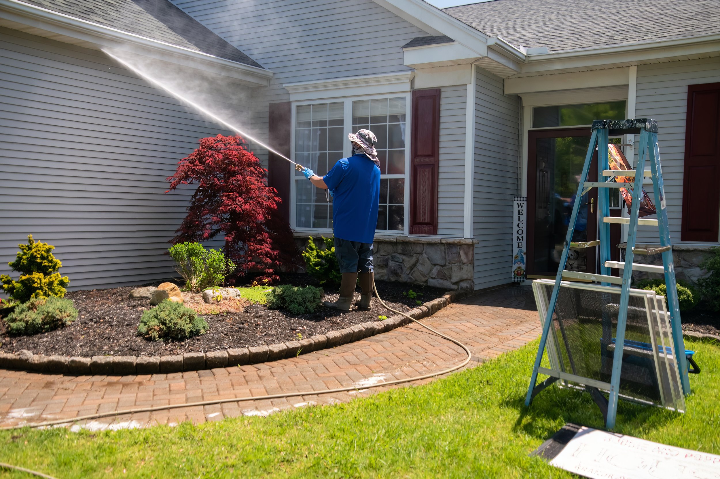 powerwashing a light grey house with burgandy shutters. Brick pavers are the pathway. Japanese maple in the flower bed. Blue ladder in the grass. Welcome sign by the front door. Cleaning gutters