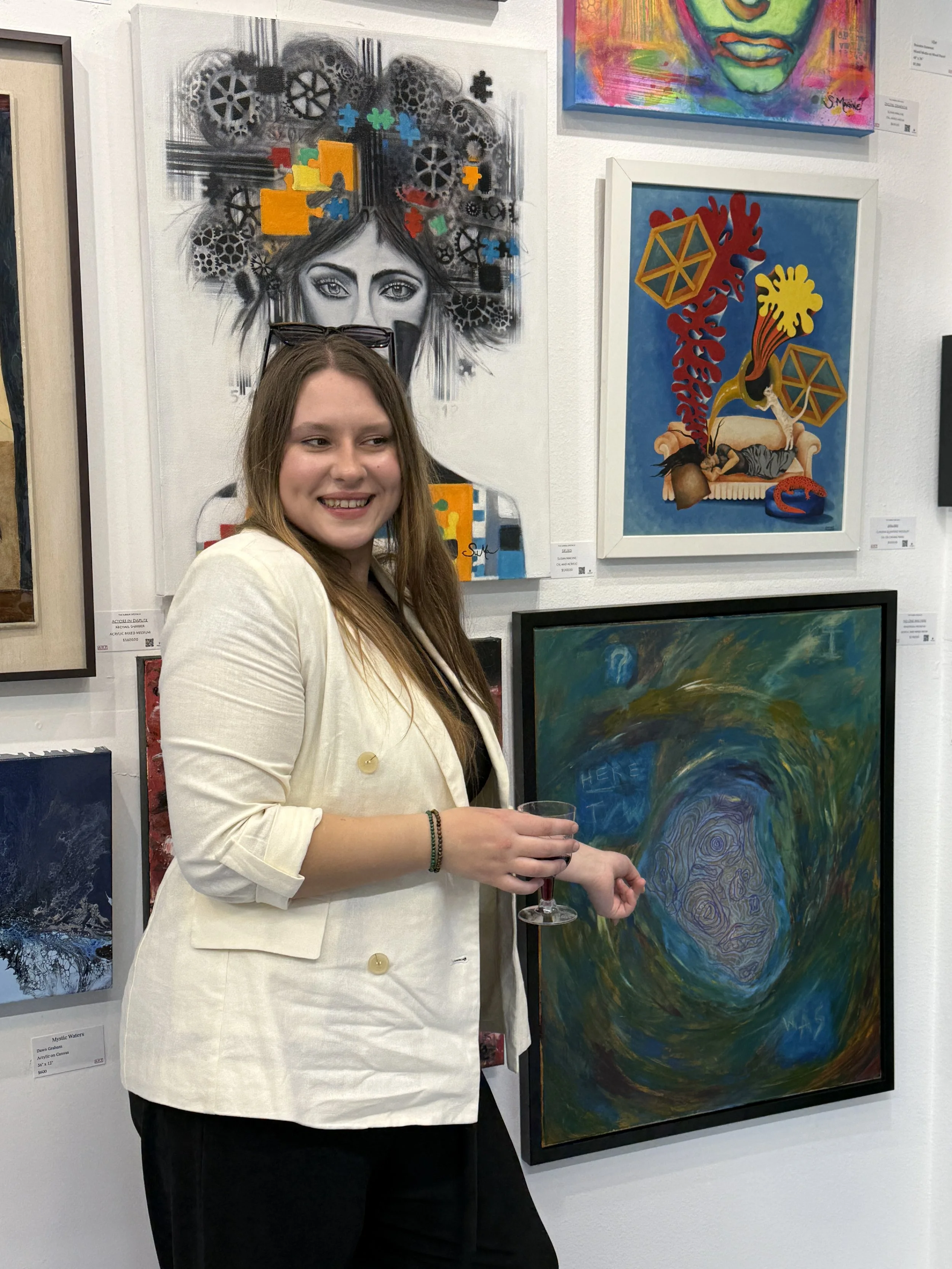 Young woman in cream blazer holding a glass of wine at an art gallery, surrounded by colorful abstract paintings and artwork.