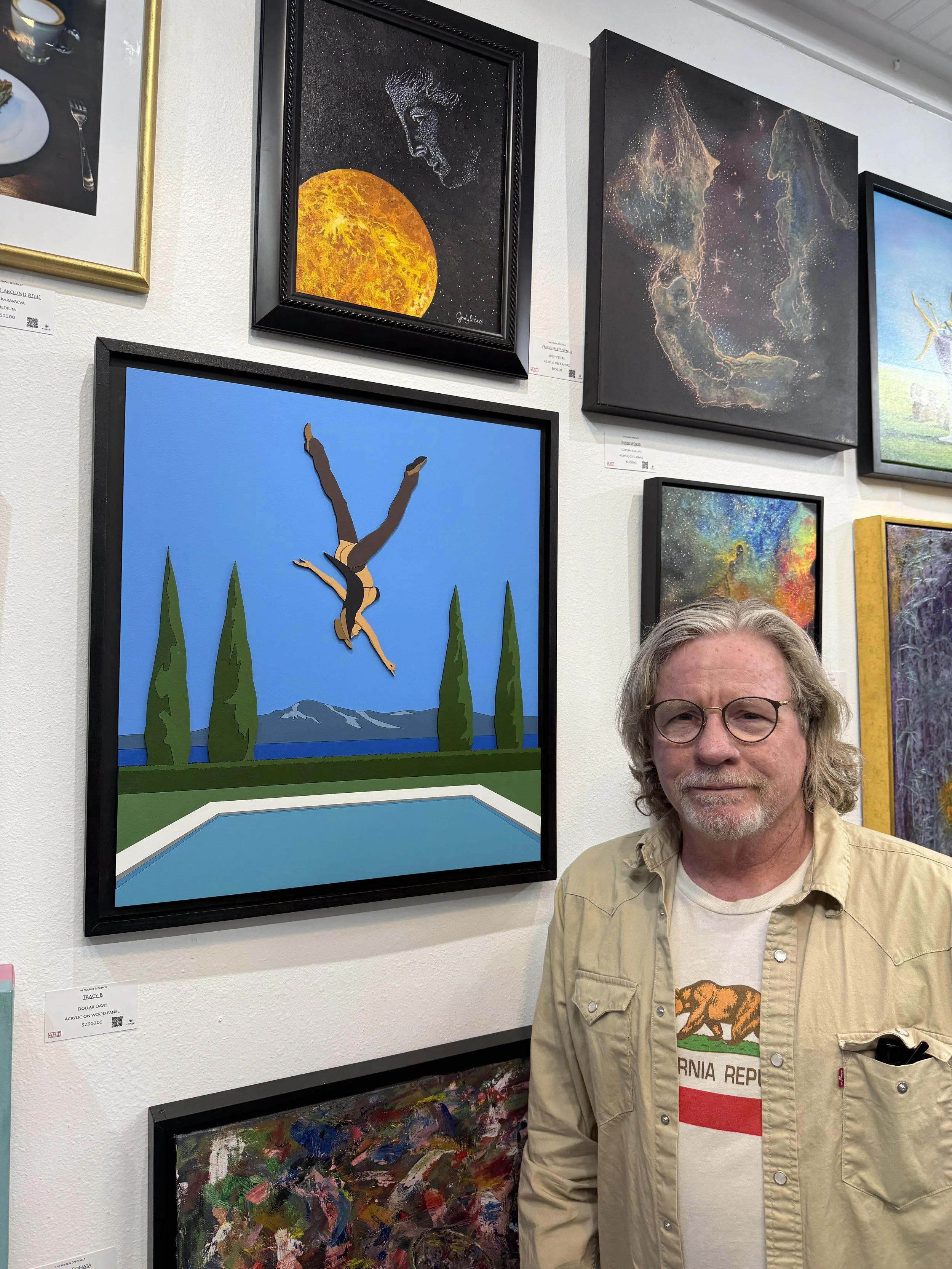 A man with gray hair and glasses standing in front of a wall with various colorful paintings, including a painting of an upside-down diver, a moon, and abstract space-themed art.