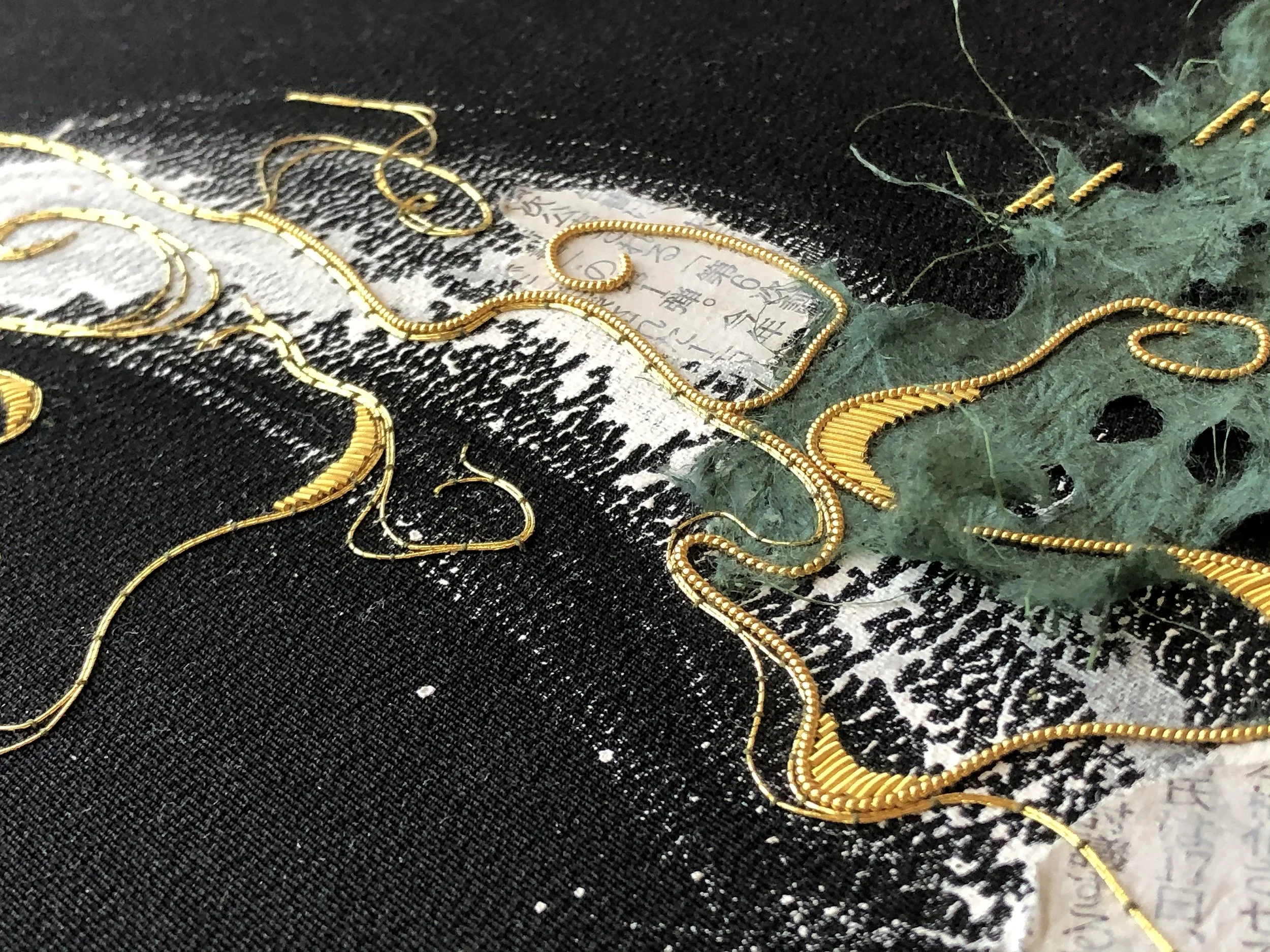 Close-up of intricate embroidery with gold threads on a black and white fabric, alongside green fabric with text.