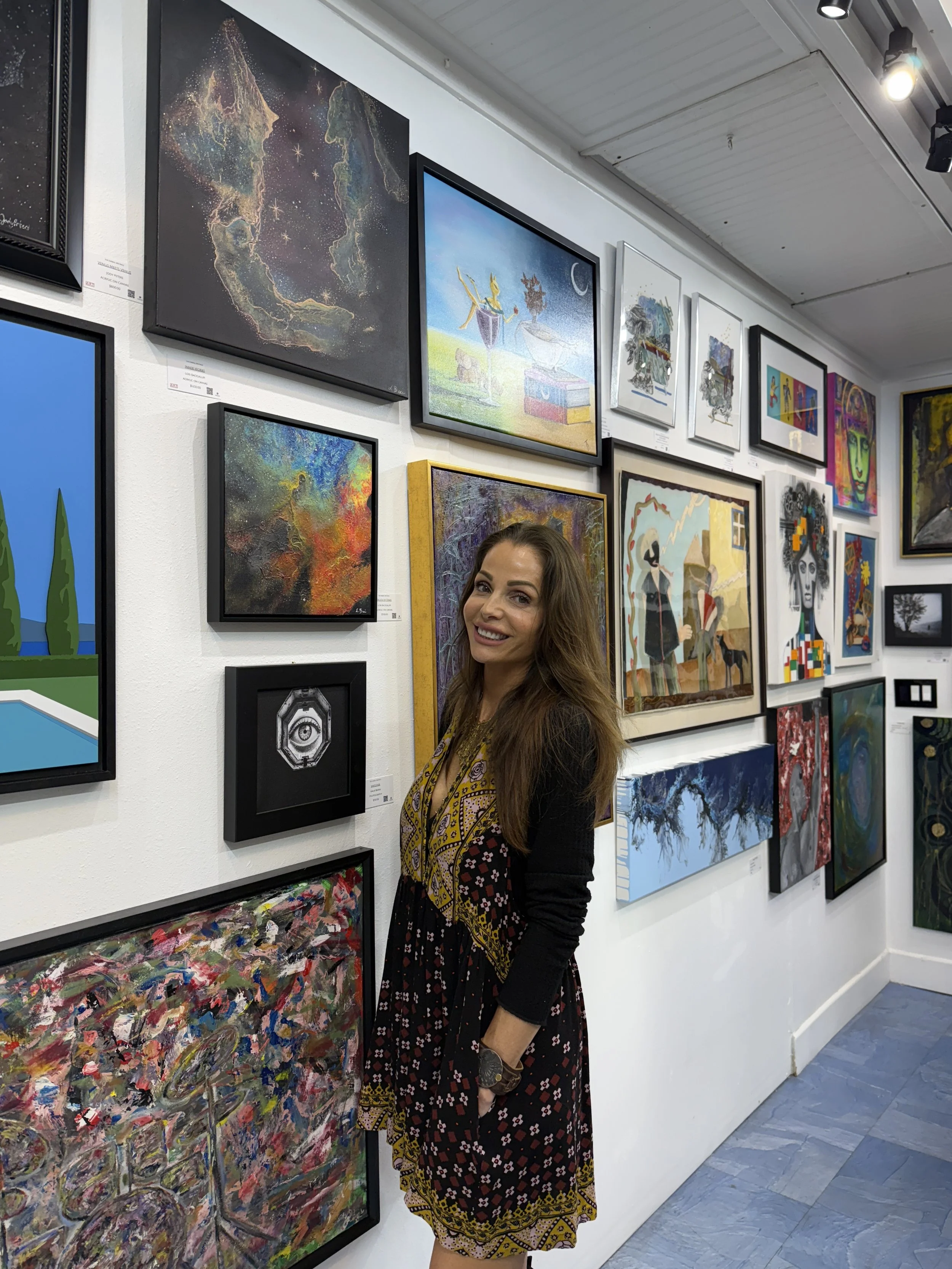 Woman with brown hair smiling at an art gallery among colorful paintings.