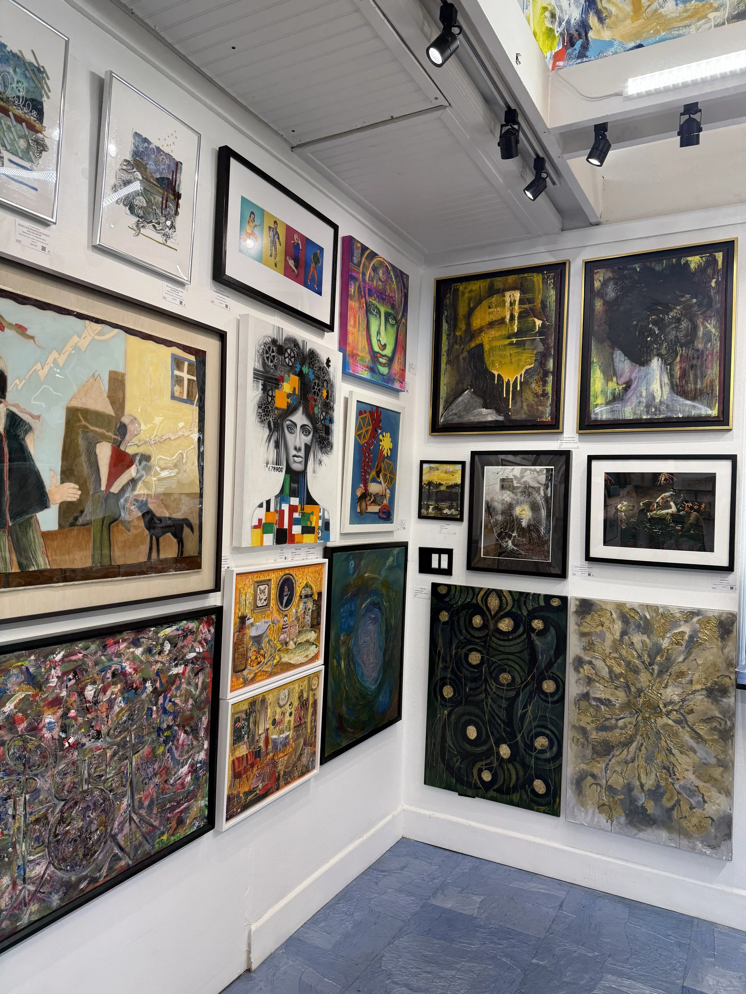 Gallery wall with various colorful and abstract paintings, including portraits, landscapes, and mixed media artwork.