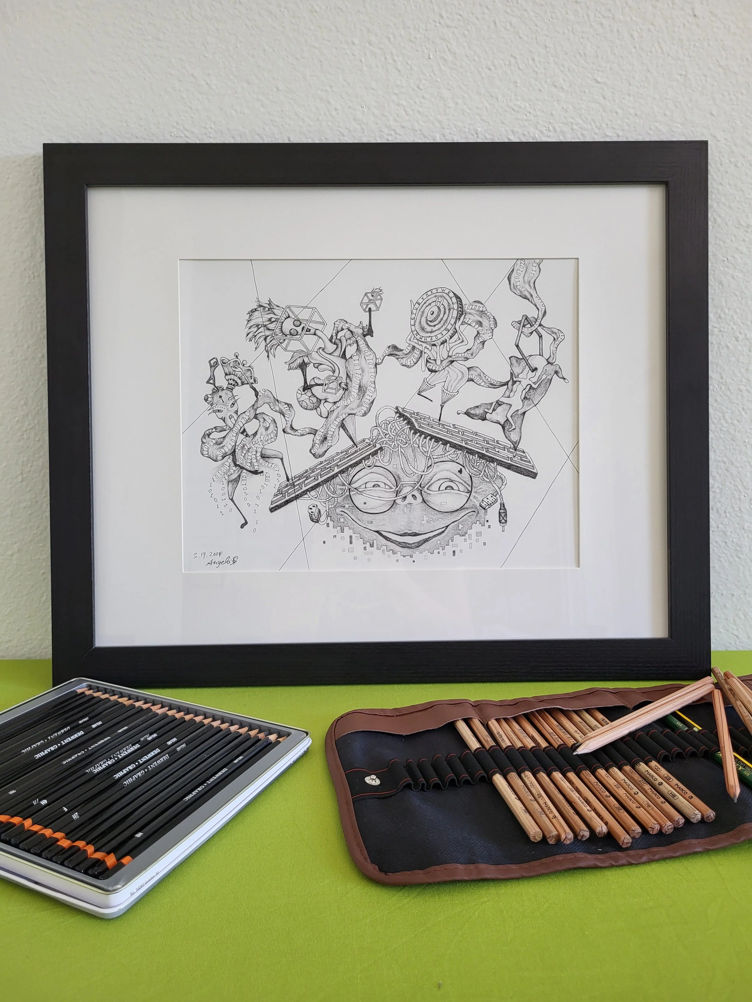 Framed black and white illustration with abstract design, featuring faces and intricate patterns, displayed on a green surface. Nearby are pencil sets in a tin case and a roll-up pouch.