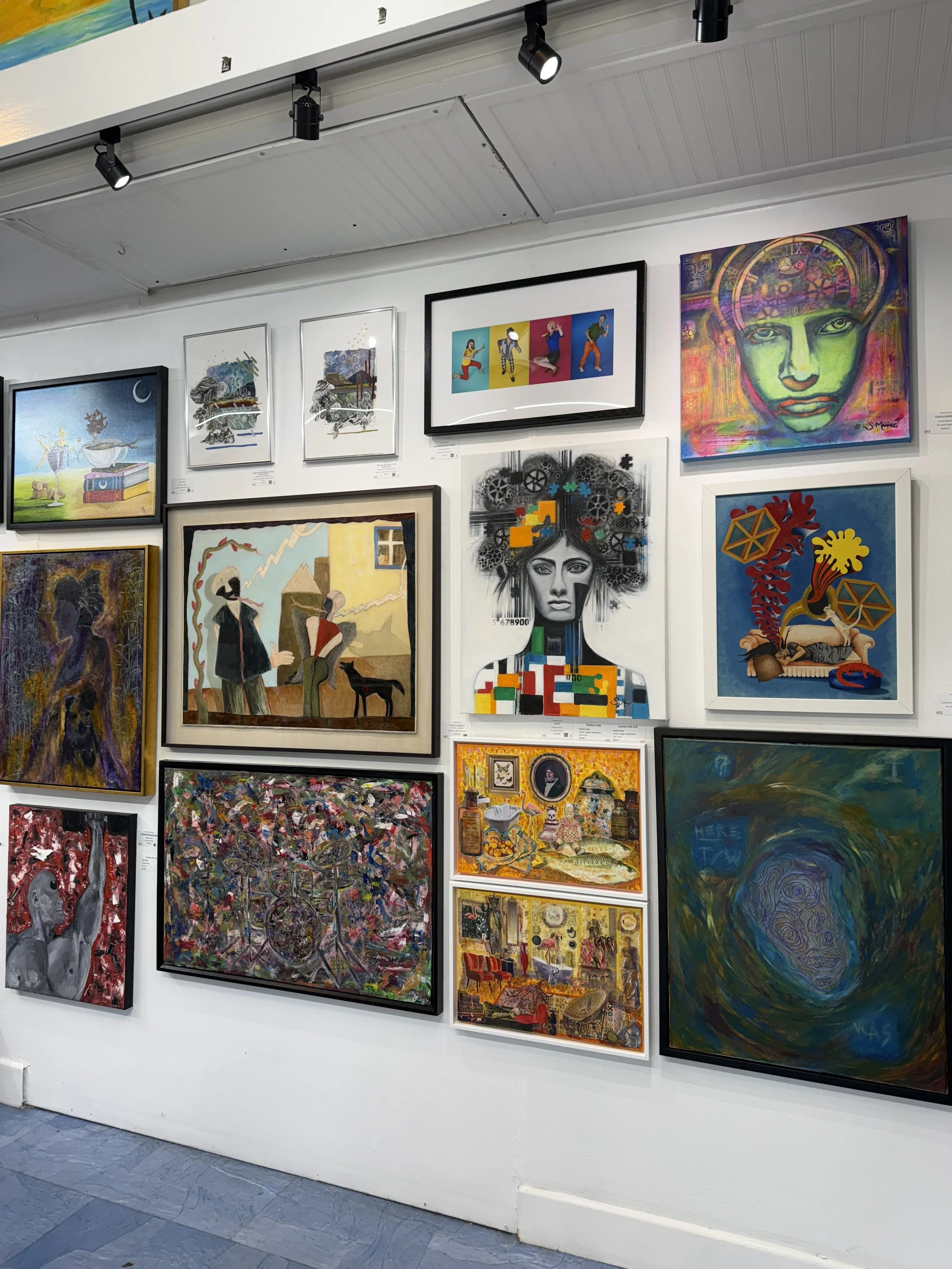 A wall in an art gallery displaying multiple colorful framed paintings, including abstract, portraits, and landscapes.
