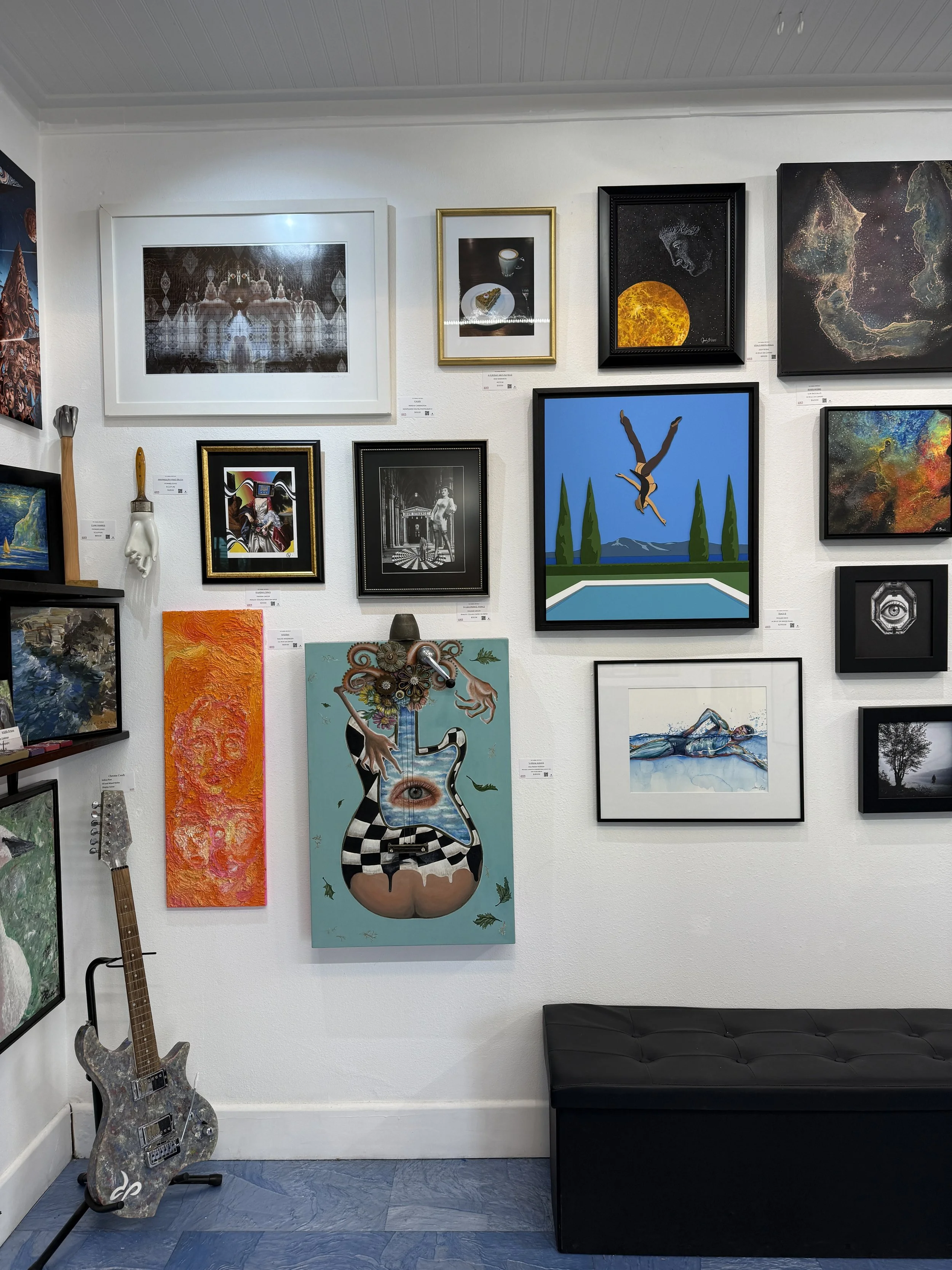 Collection of various framed paintings and photographs on a gallery wall, with a painted guitar leaning against the wall on the left and a black bench below.