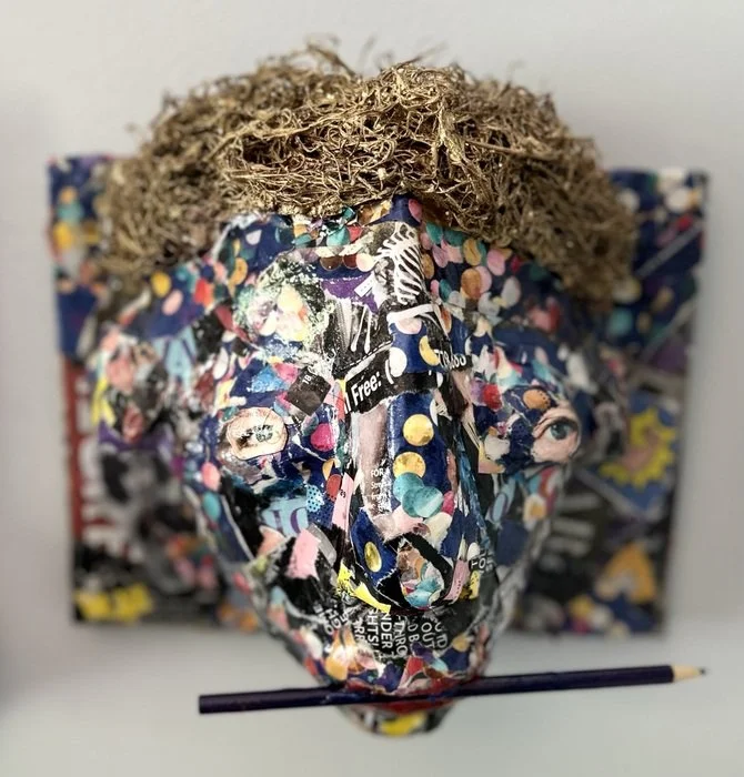 Mixed-media art piece resembling a human head with curly hair made of textured material and a face constructed from torn, colorful paper with various patterns and images, holding a pencil.