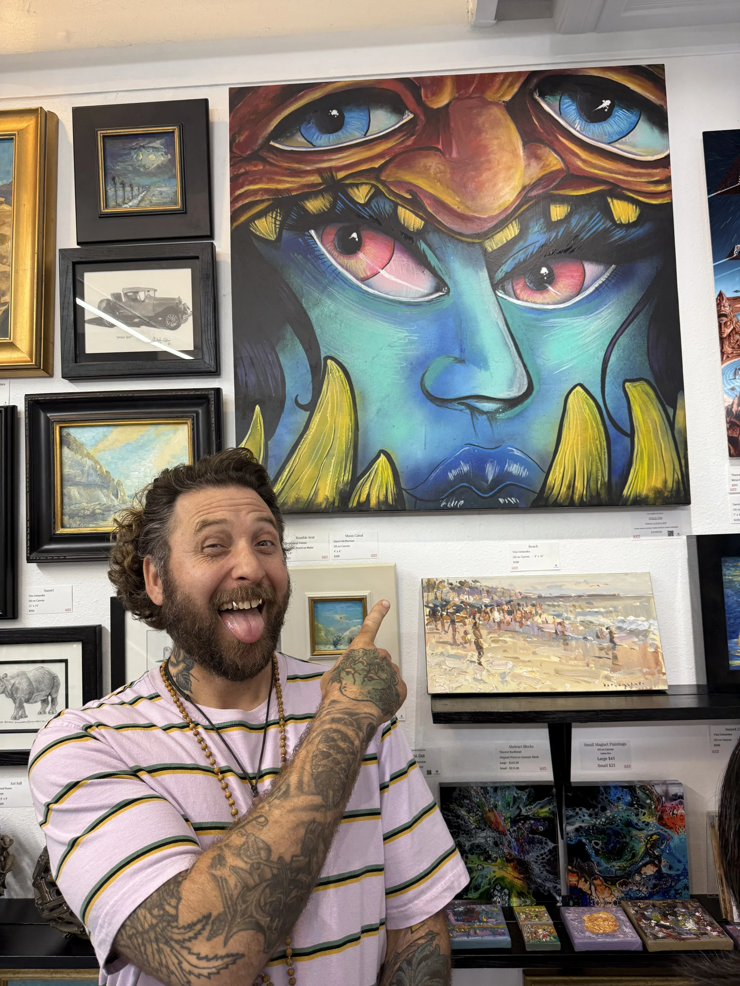A man with a beard, tattoos, and a striped pink, white, and yellow shirt is smiling, sticking out his tongue, and pointing at a vibrant, surreal painting of a face with multiple eyes, blue skin, and yellow claws on a gallery wall.