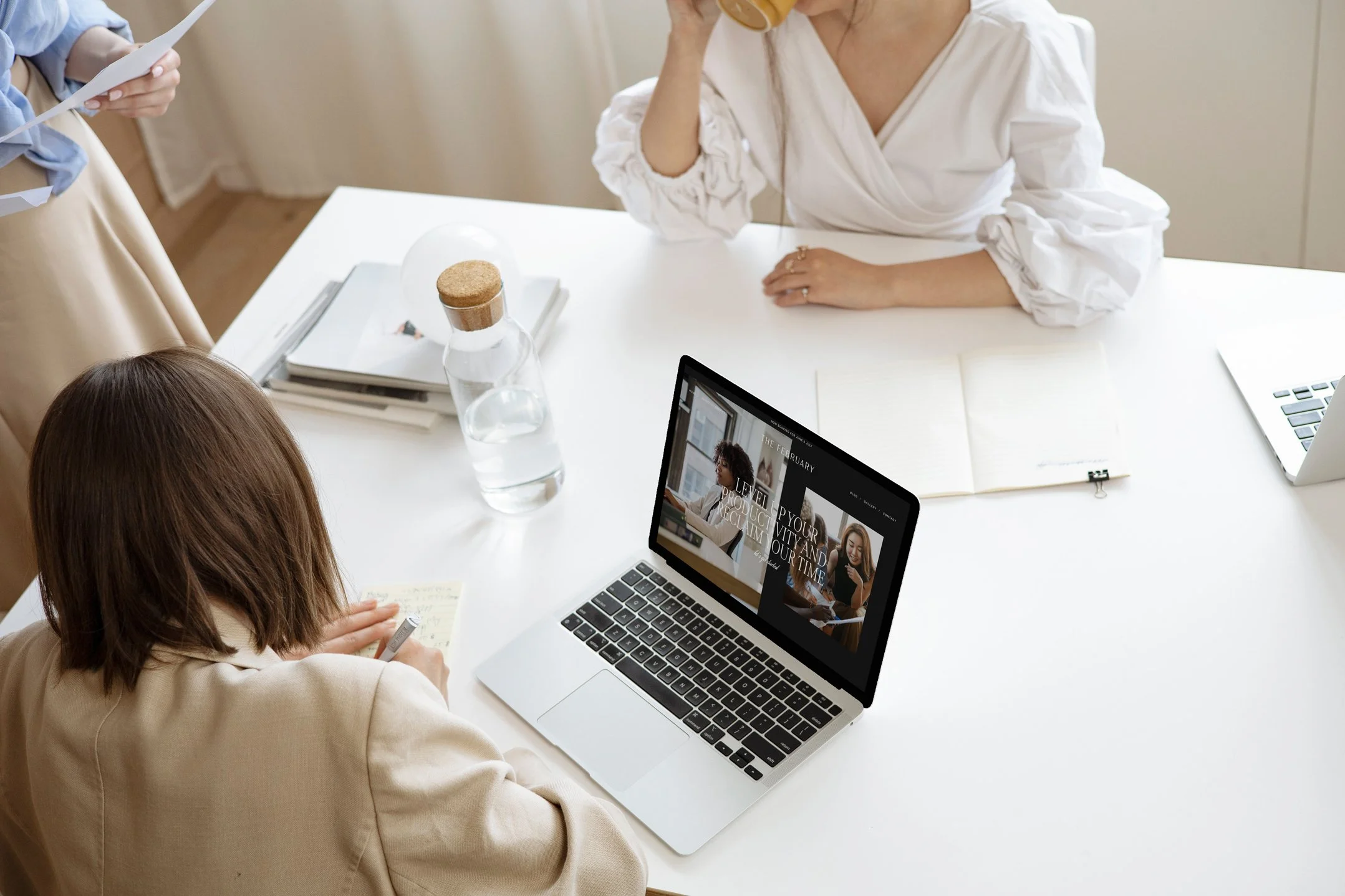 Two women in a meeting room, one taking notes and the other drinking coffee, with a laptop displaying a website, a notebook, and water on the table.