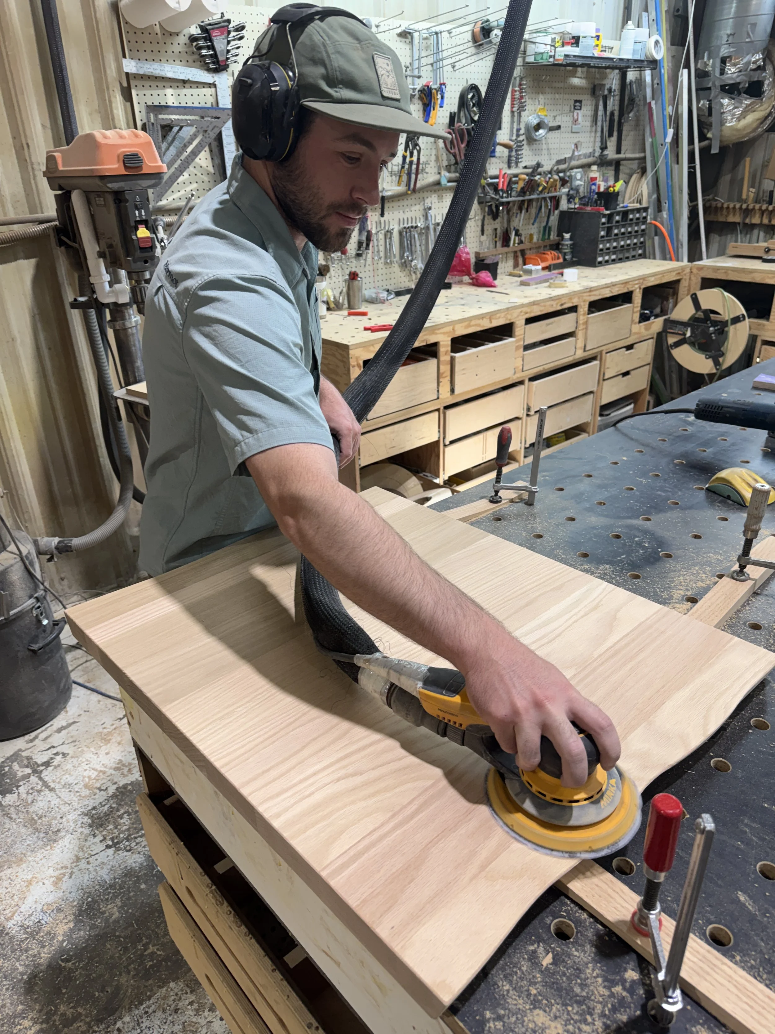 Man sanding a wooden piece in a woodworking shop with tools and shelves in the background.