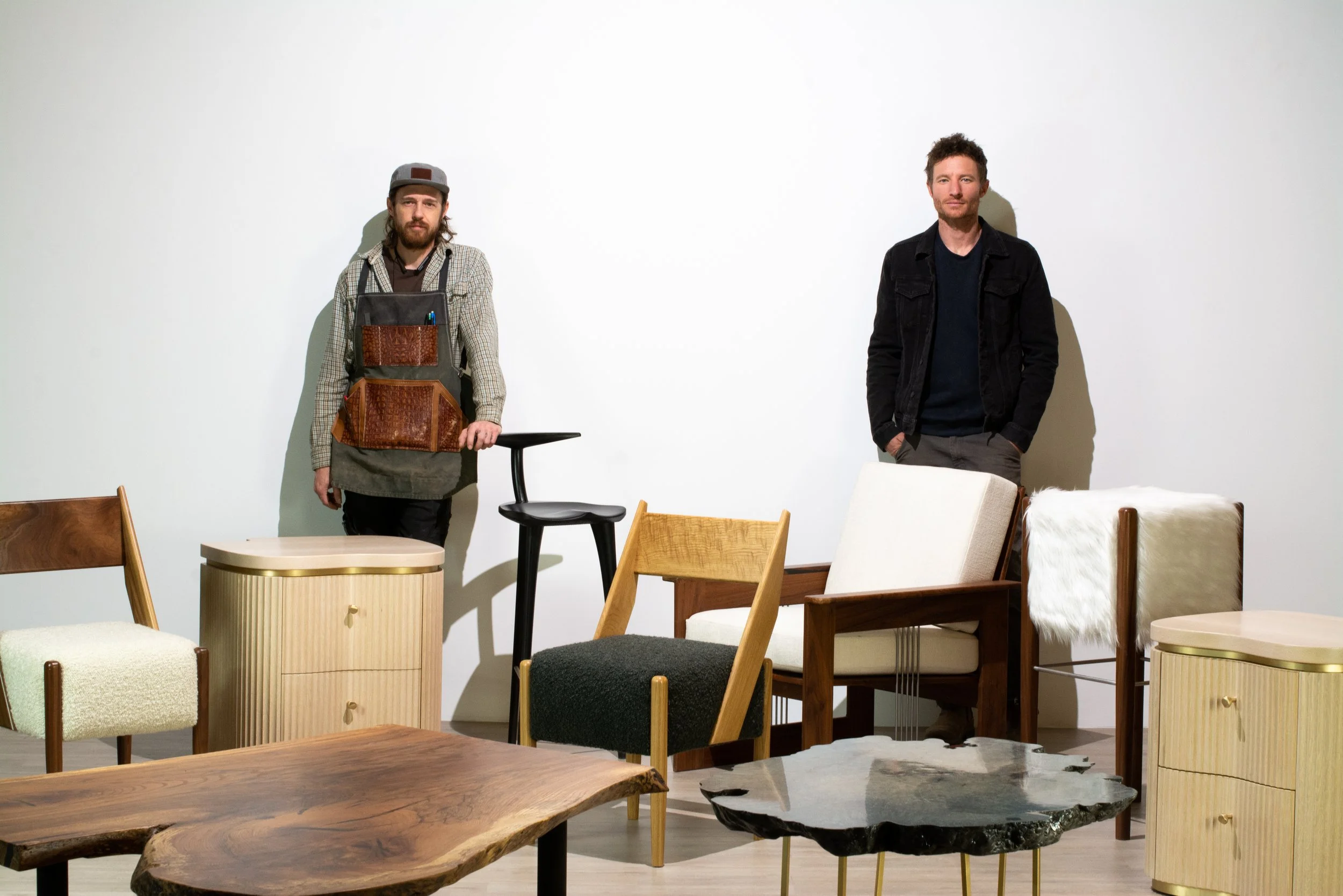 Two men standing in a room with wooden and upholstered chairs and tables, one wearing a checkered shirt and apron with a hat, the other in a black jacket, against a plain white wall.