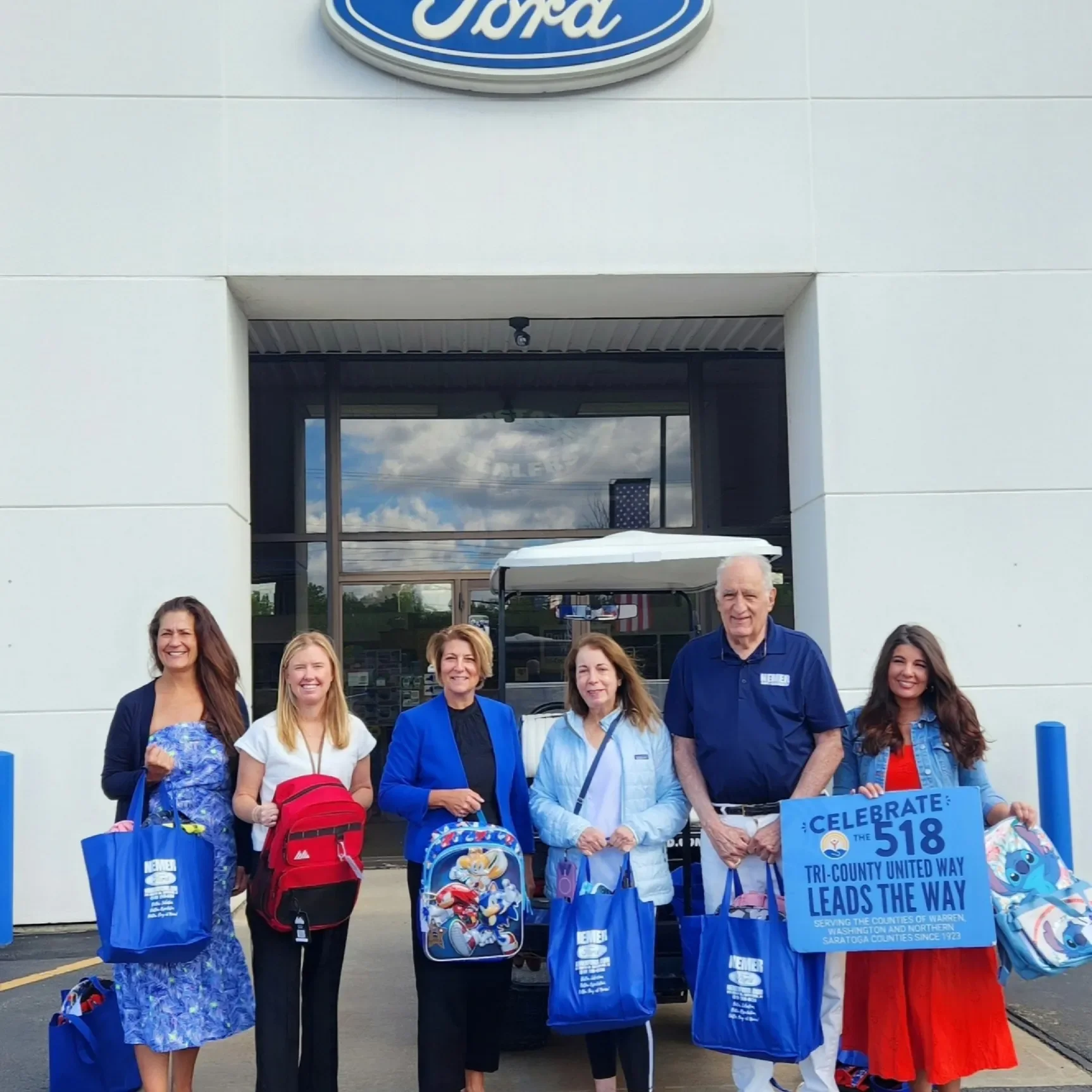 A group of people standing outside a building with a Ford logo, holding bags and a sign that reads 'Celebrate the 518 Tri-County United Way Leads the Way.' The group seems to be participating in an event related to the United Way.