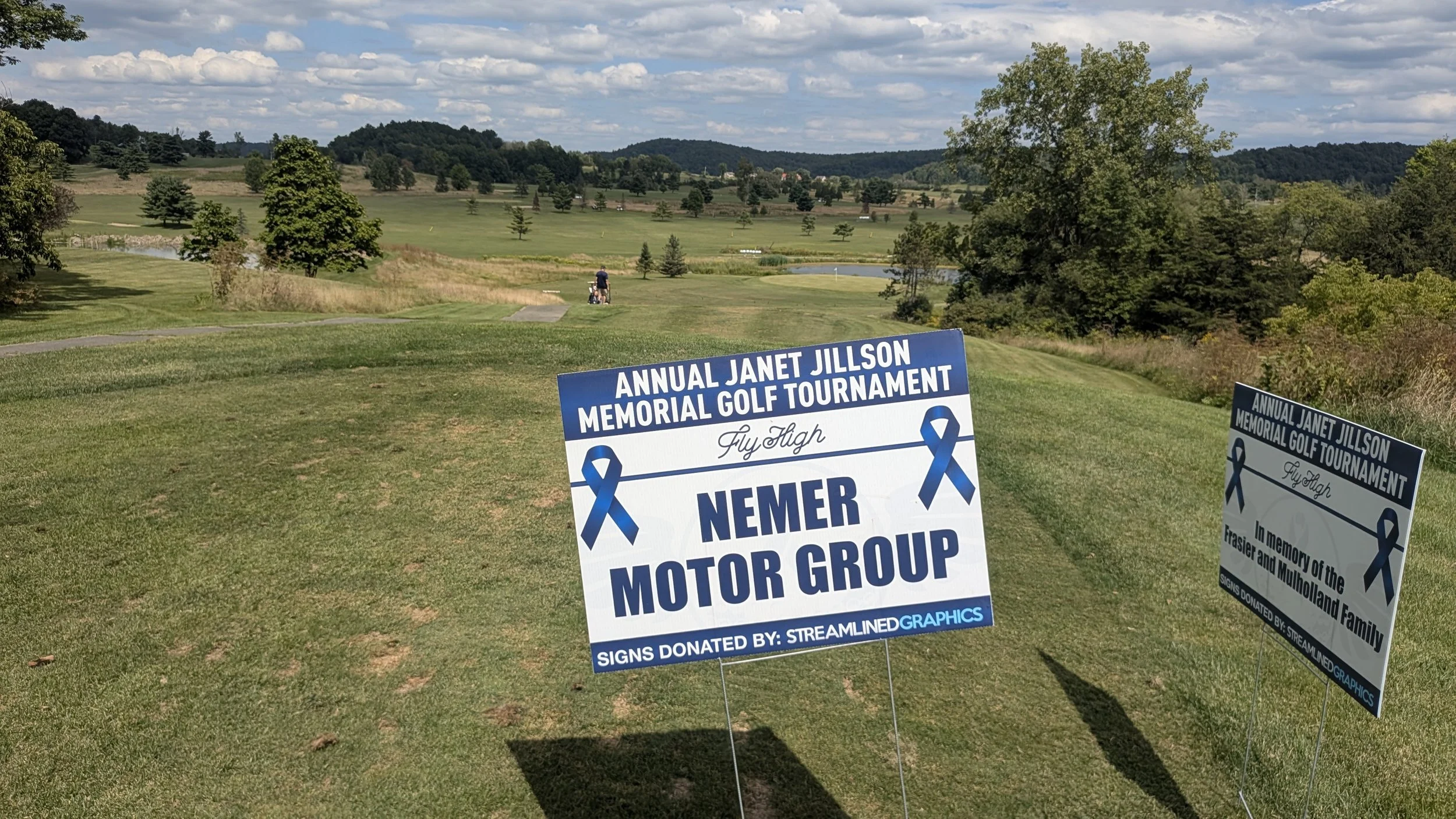 A scenic view of a golf course with green fairways, trees, and hills in the background under a partly cloudy sky. Two signs in the foreground mention the Annual Janet Jillson Memorial Golf Tournament, with ribbons and logos, and a person is walking near a golf cart in the distance.