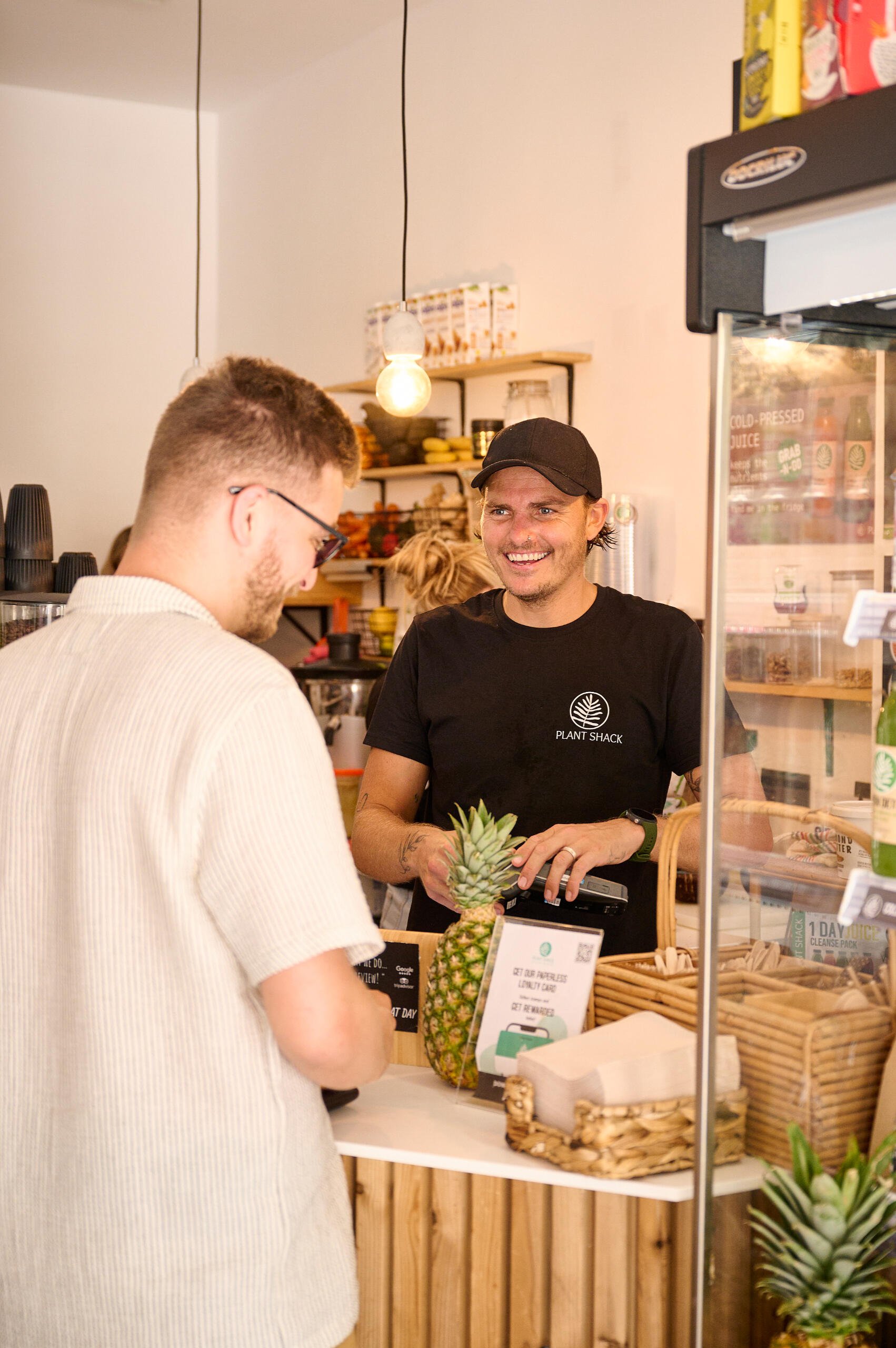 A smiling cashier wearing a Plant Shack t-shirt and a black cap serves a customer at a juice shop counter. The customer wears glasses and a beige shirt, with a pineapple on the counter. The shop has shelves with yellow and black items and hanging light bulbs.