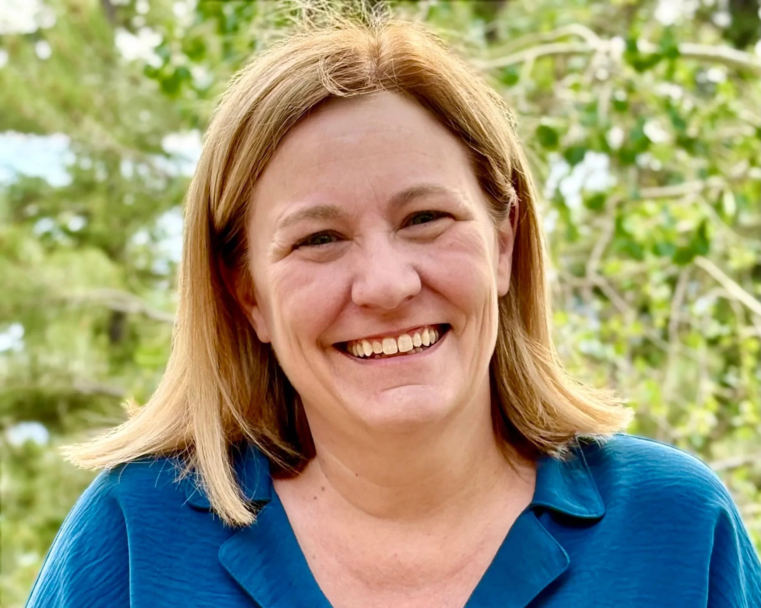 A smiling woman with blonde hair wearing a blue shirt outdoors with trees in the background.