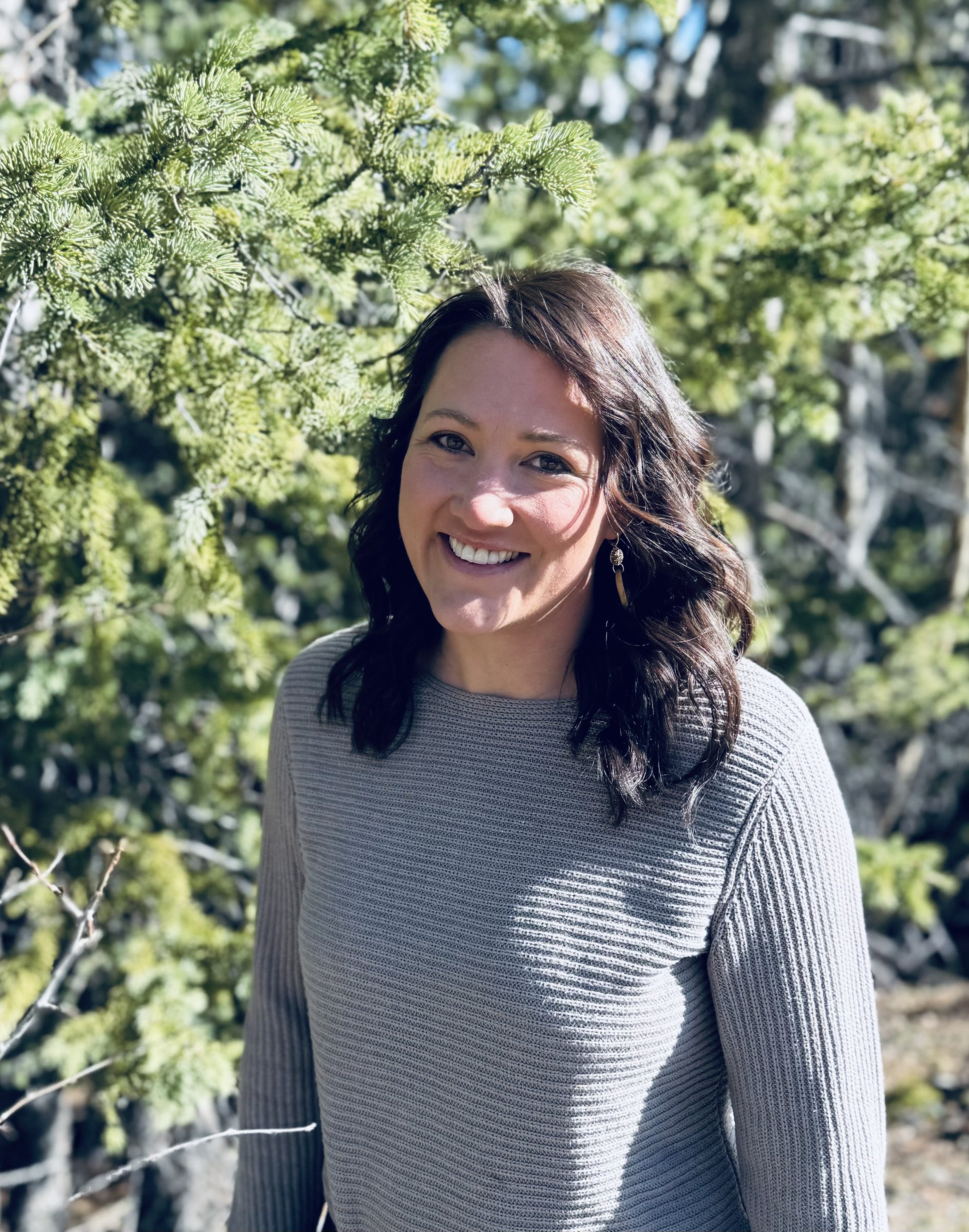 A woman with long dark hair and earrings smiling in an outdoor setting with green tree branches and blue sky in the background.