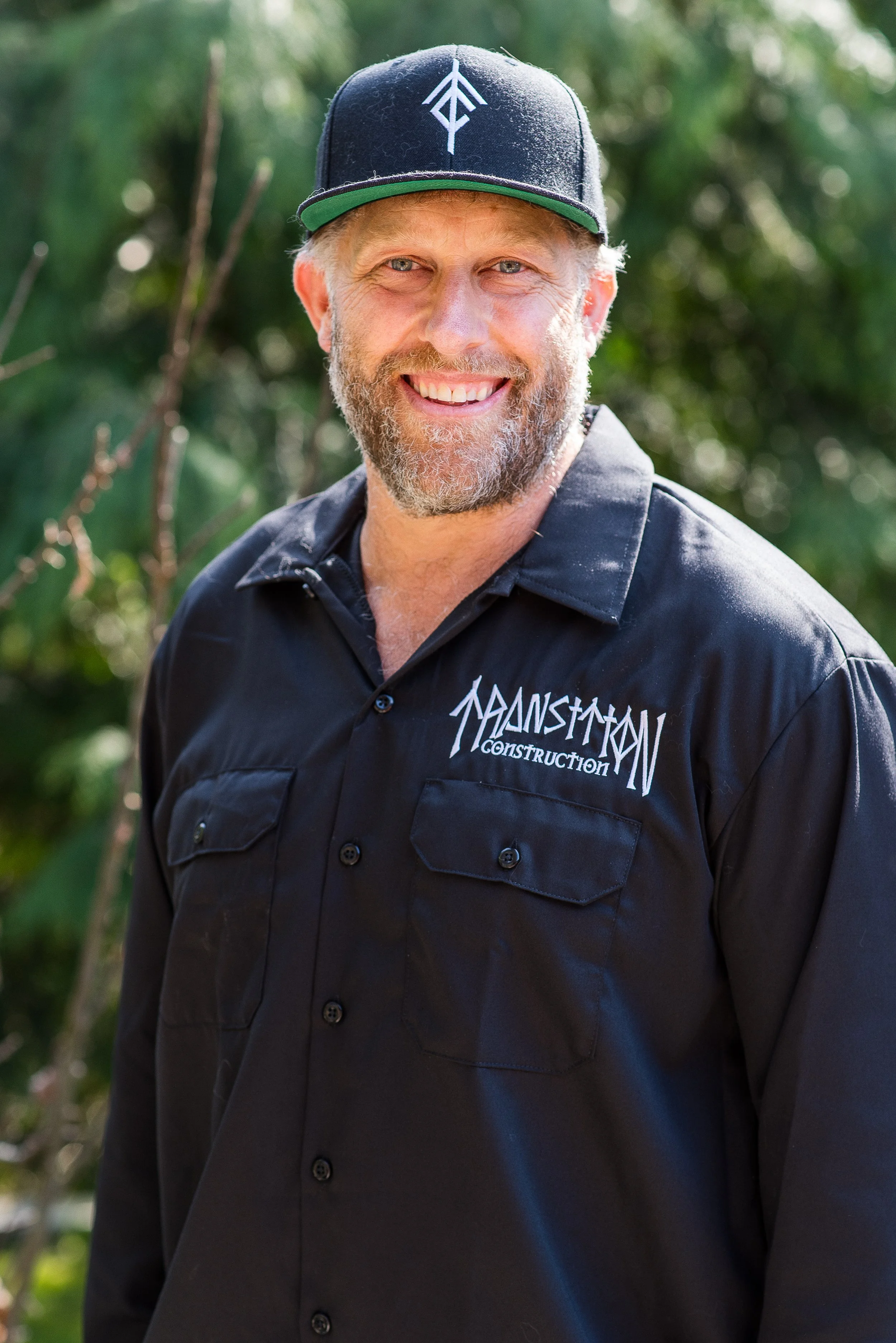 A smiling man with a beard and blue eyes, wearing a black baseball cap and a black work shirt with 'Mainstream Construction' embroidered on it, outdoors with green foliage in the background.