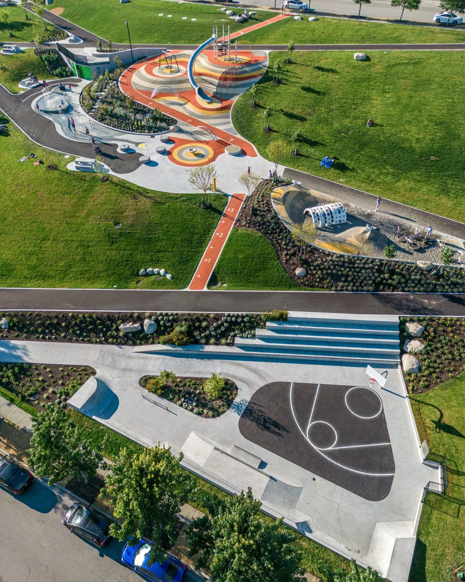 Aerial view of a colorful public playground with slides, climbing structures, and seating areas, surrounded by green lawns and trees. Sheffield park in Port Coquitlam