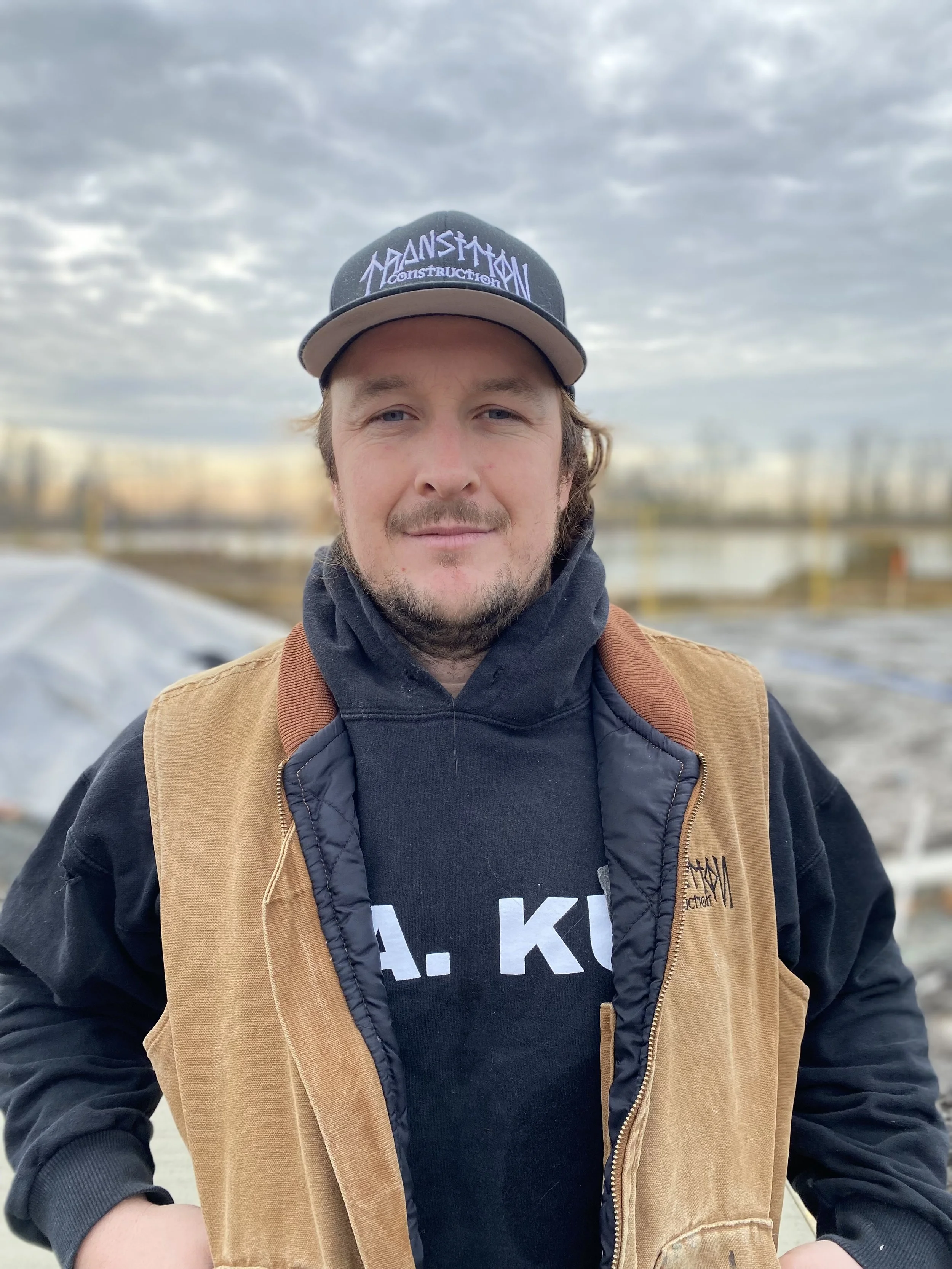 A man wearing a black baseball cap, black hoodie, and tan vest, standing outdoors with a cloudy sky and industrial area in the background.