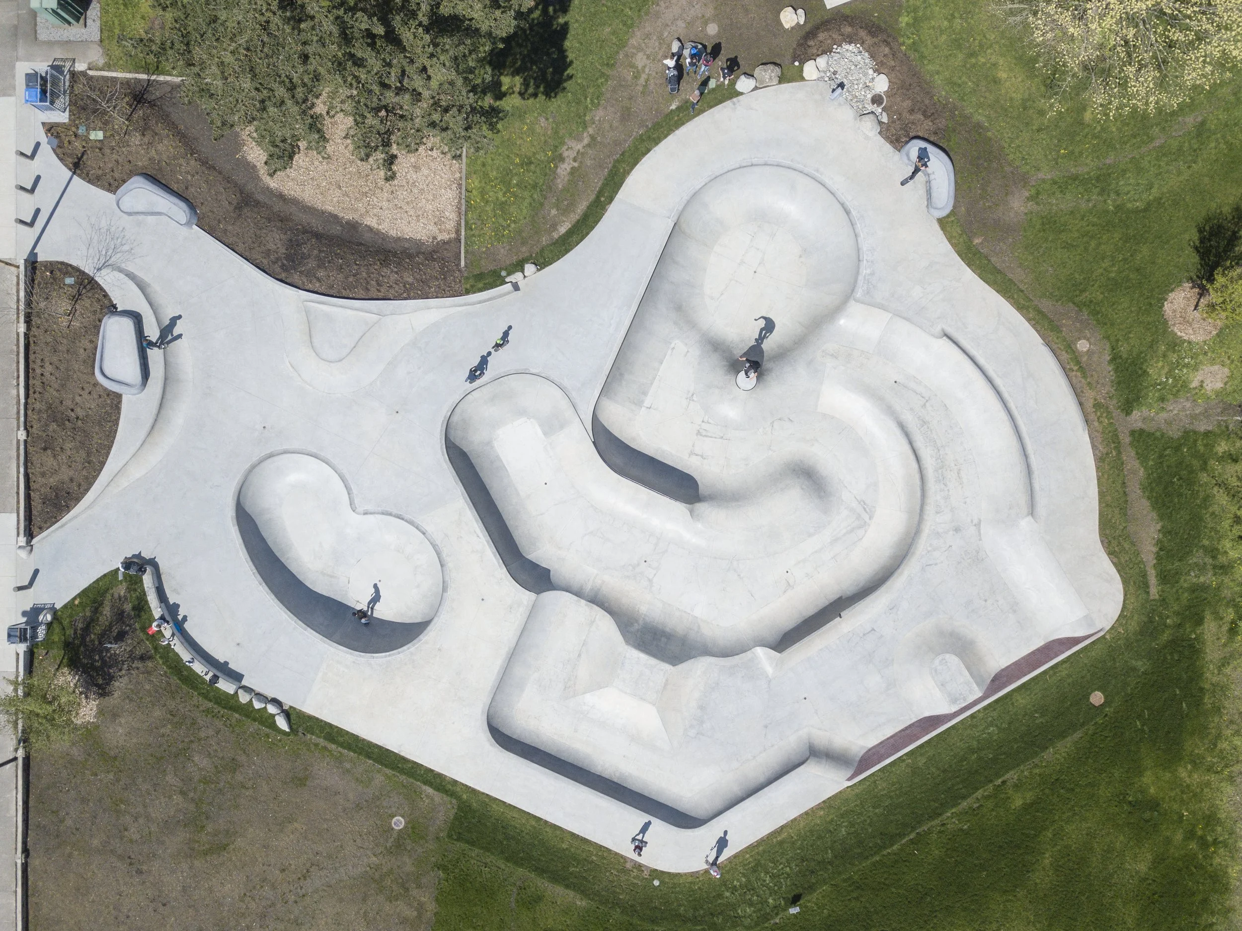 Aerial view of a skate park with various concrete ramps and bowls, surrounded by grass and trees, with several people skateboarding and riding bikes.