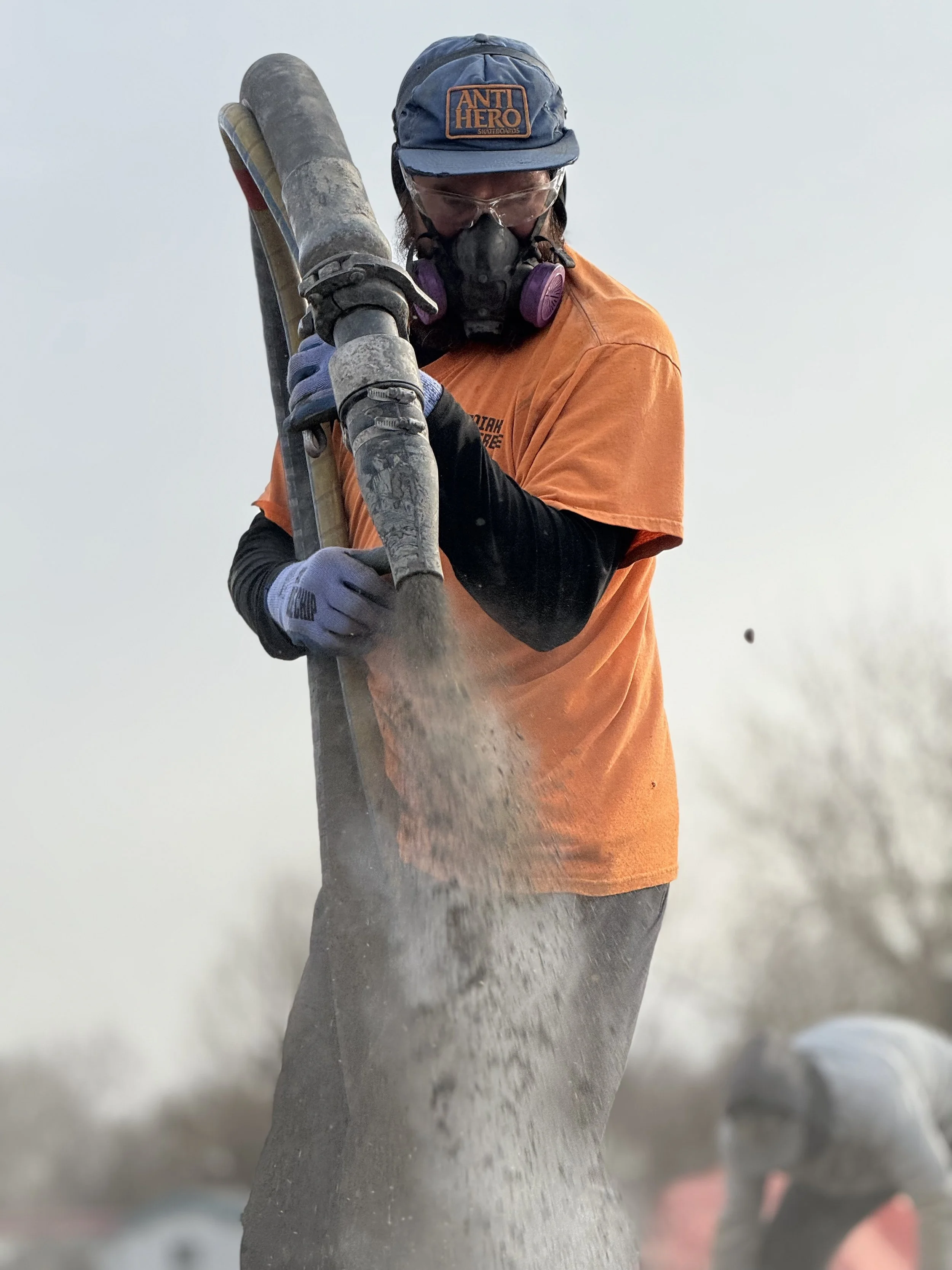 Person wearing an orange shirt, black gloves, a mask, and a blue cap labeled 'ANTI HERO,' operating a  Shot Crete  concrete nossle