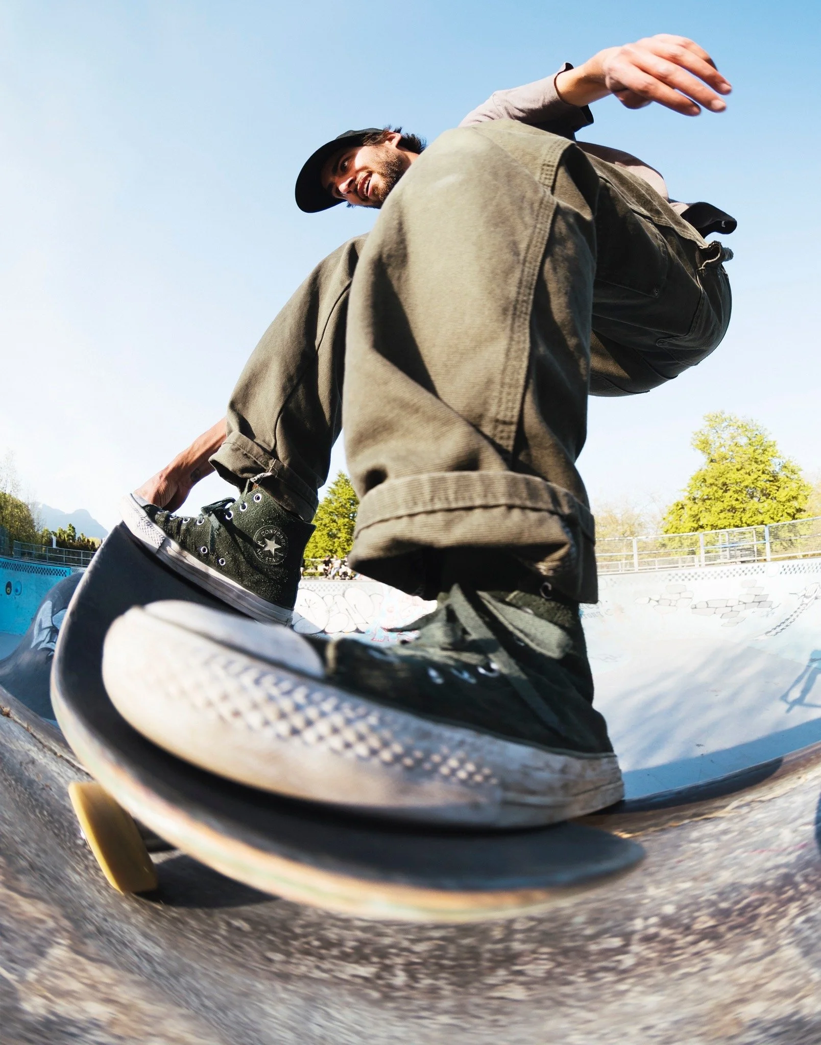 Skateboarder in mid-ride at a skate park during daytime, wearing black sneakers, khaki jacket, and a black cap, with a smile on his face, surrounded by trees and a blue sky.
