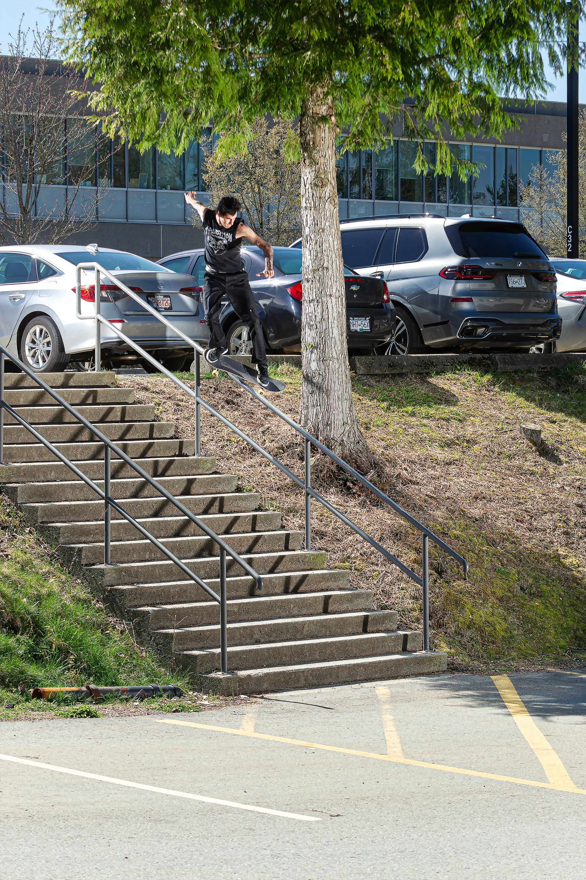 Skateboarder performing a trick on a handrail beside a staircase outside a parking lot.