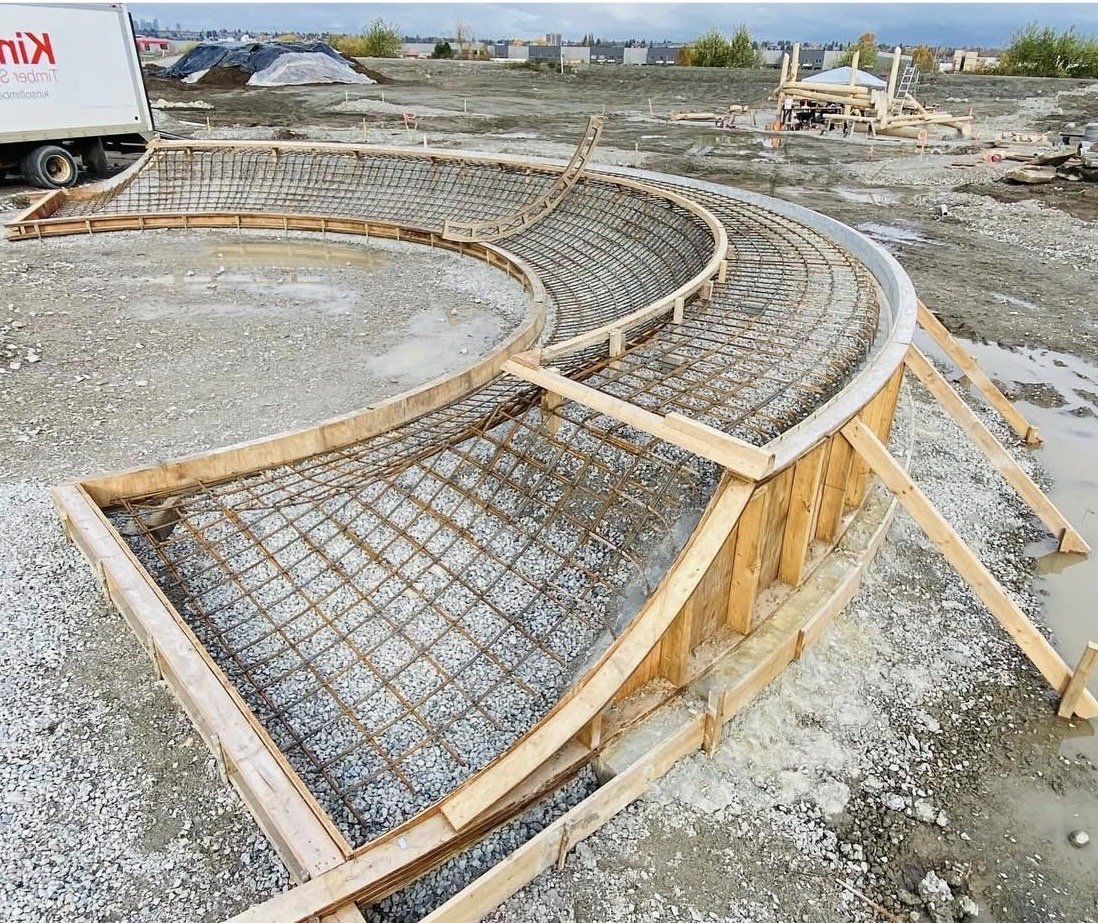 Construction site with a wooden curved formwork and steel reinforcement for a concrete structure, with a construction truck nearby.