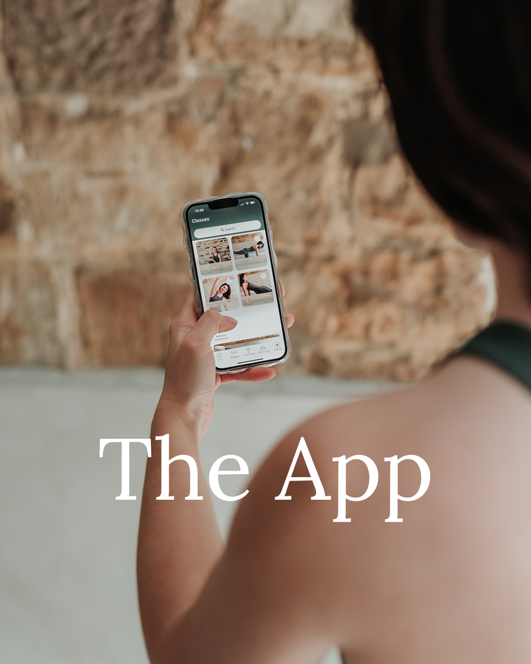 Person holding a smartphone displaying a fitness app with workout class images, standing in front of a brick wall, with text overlay 'The App'.
