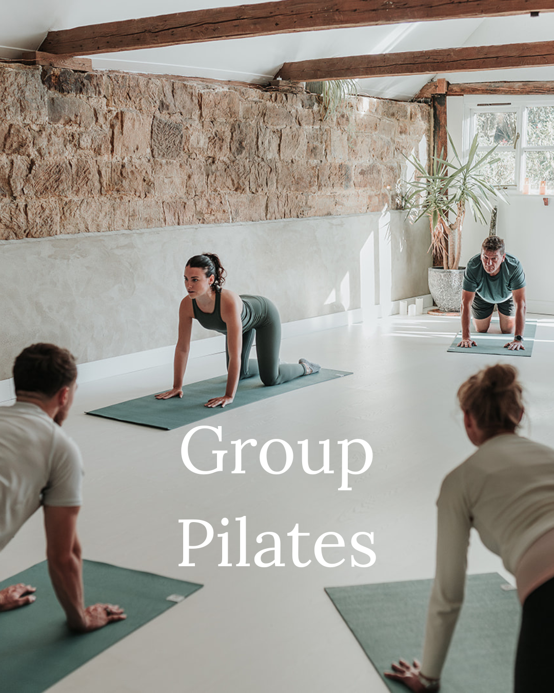 A group of four people participating in a Pilates class in a bright studio with a stone wall and large window, with women and men on yoga mats practicing Pilates exercises.
