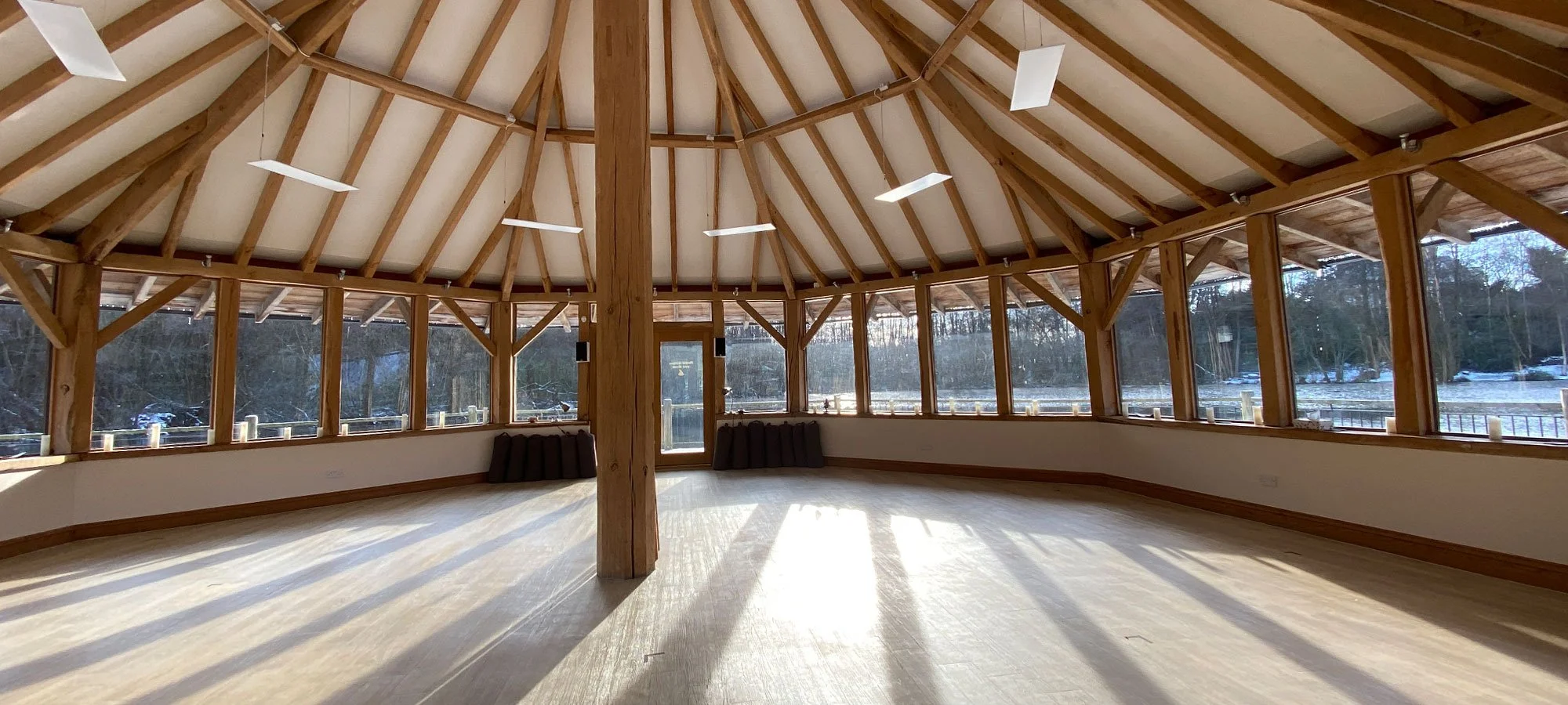 Empty room with wooden beams and curved ceiling, surrounded by large windows showing a snowy outdoor landscape with trees and water, sunlight streaming in.