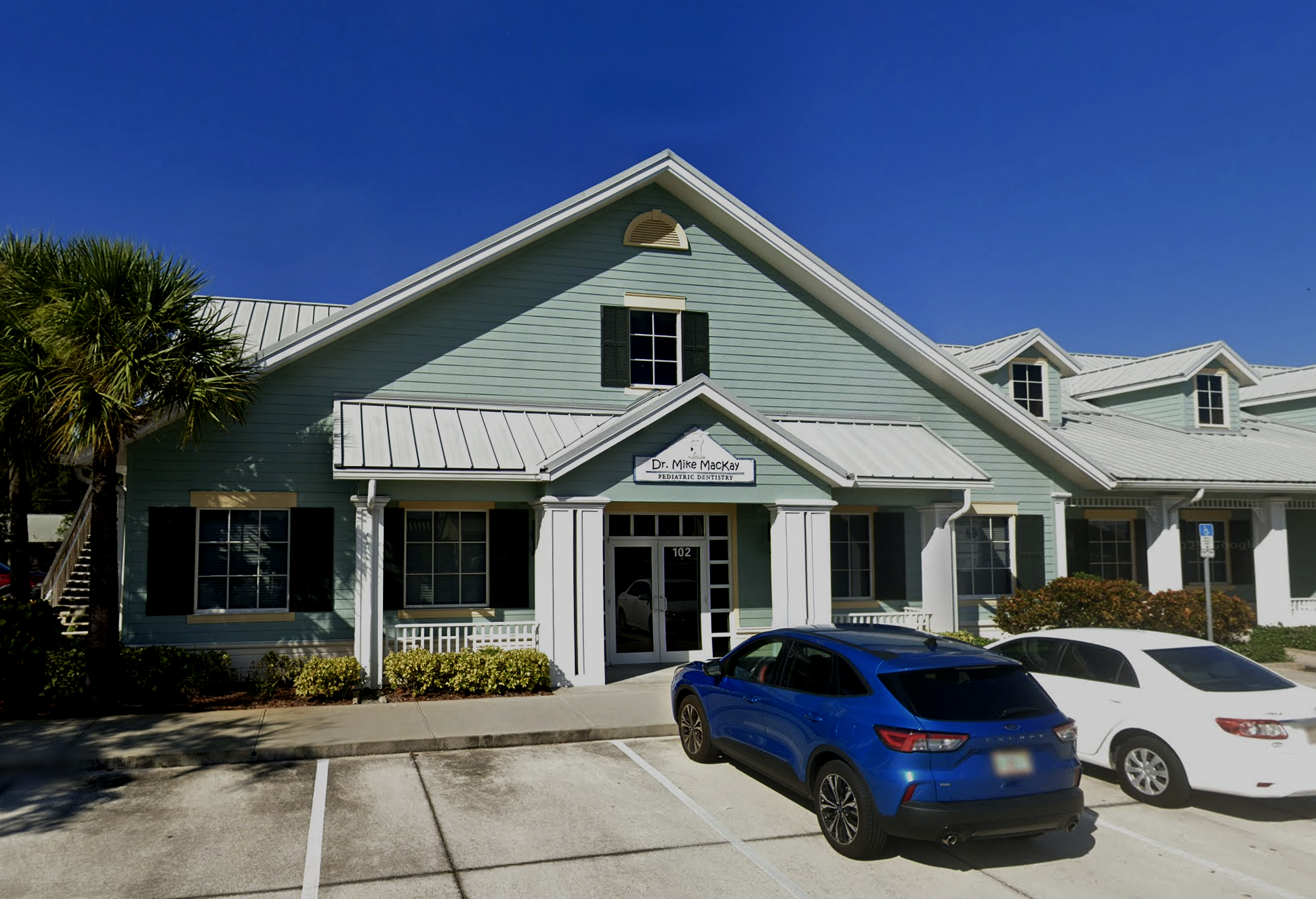 A two-story pastel blue building with white trim, labeled as Dr. Mike MacKay Pediatric Dentistry, with a small parking lot in front and several parked cars, including a blue car and a gray car. The building has black window shutters and a metal roof, with palm trees on the left side.