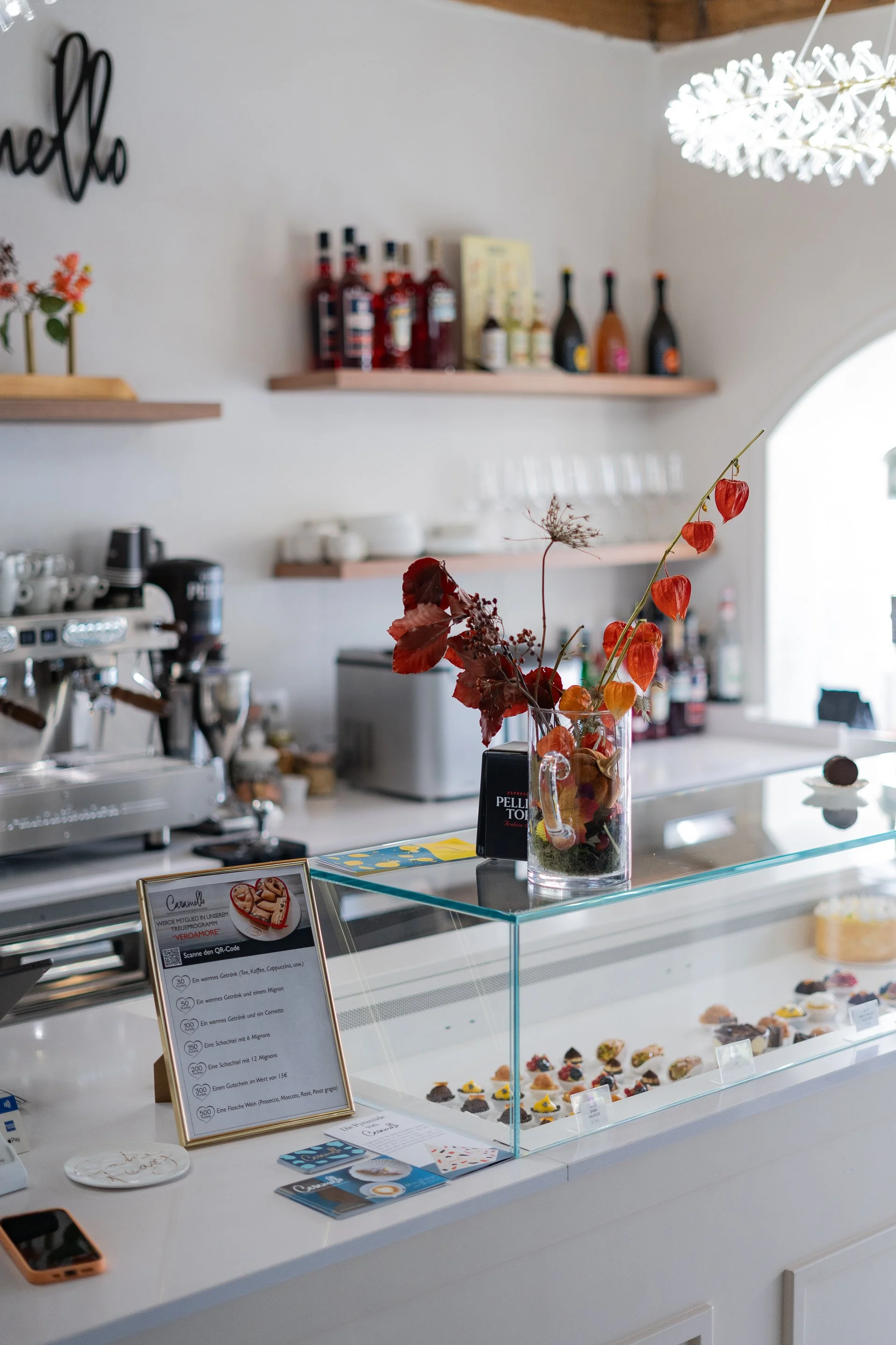 Inneneinrichtung eines Cafés mit einer Kuchentheke, die verschiedene kleine gebackene Süßigkeiten zeigt, dekorierten Blumen in einer Vase und einem Menü. Im Hintergrund sind Regale mit Flaschen und Geschirr sowie eine Espressomaschine sichtbar.