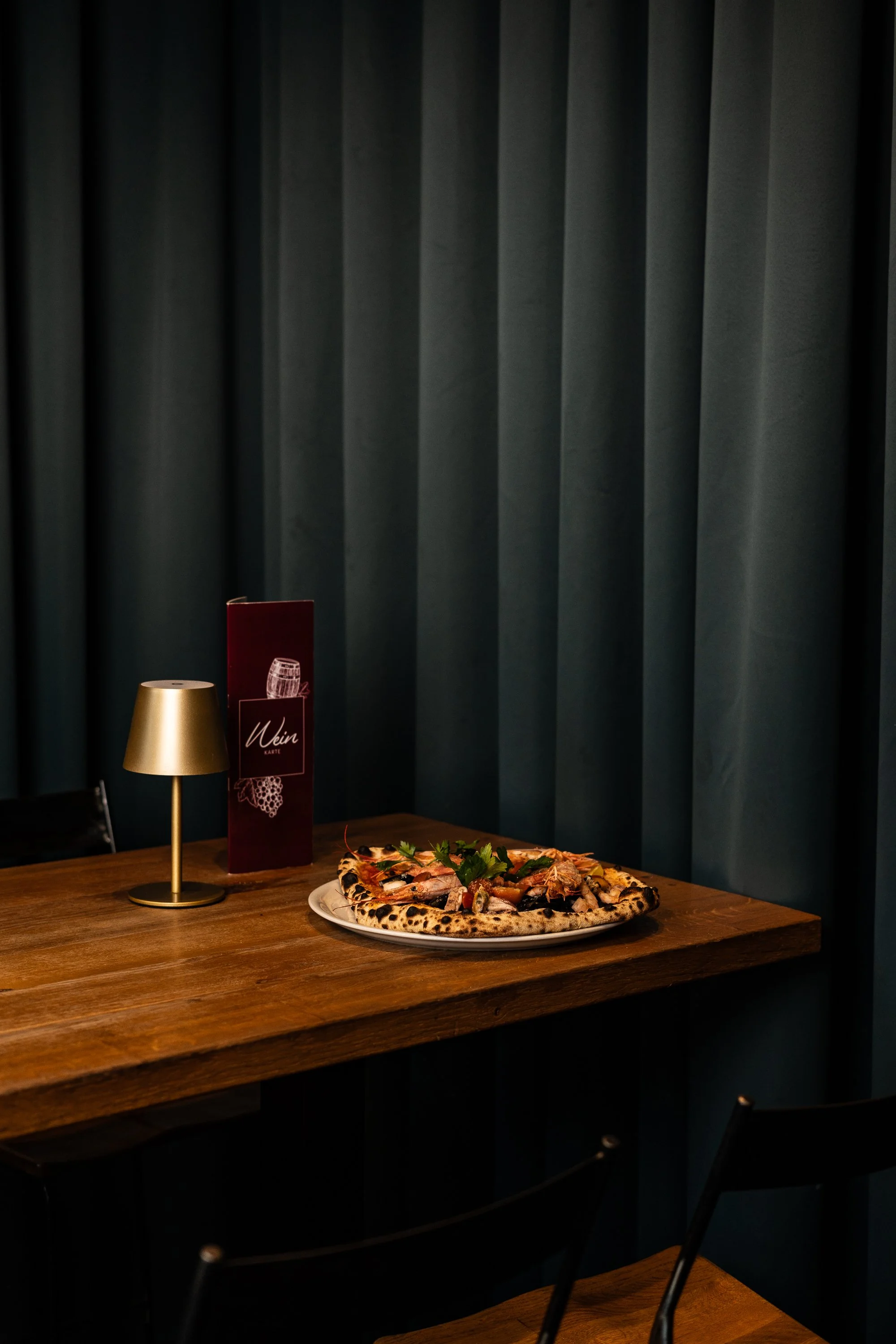 Pizza auf einem weißen Teller auf einem Holztisch, mit einem kleinen Lampen und einer Weinkarte im Hintergrund vor dunklen Vorhängen.