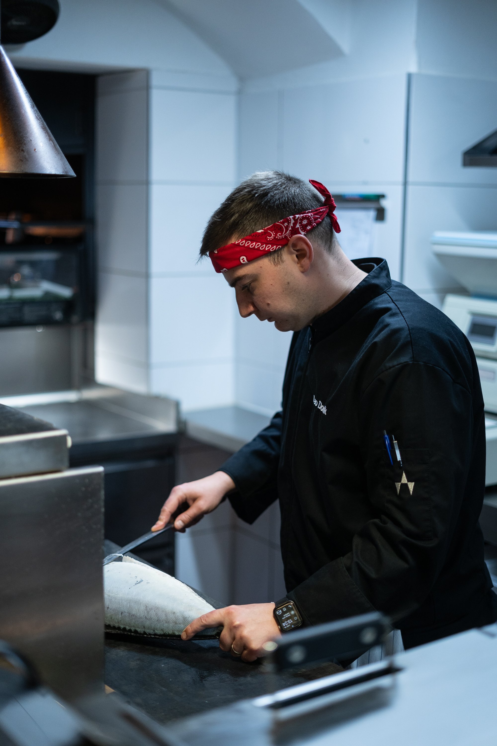 Koch beim Zubereiten eines Fischs in einer professionellen Küche, trägt einen roten Kopftuch und schwarze Kochjacke.
