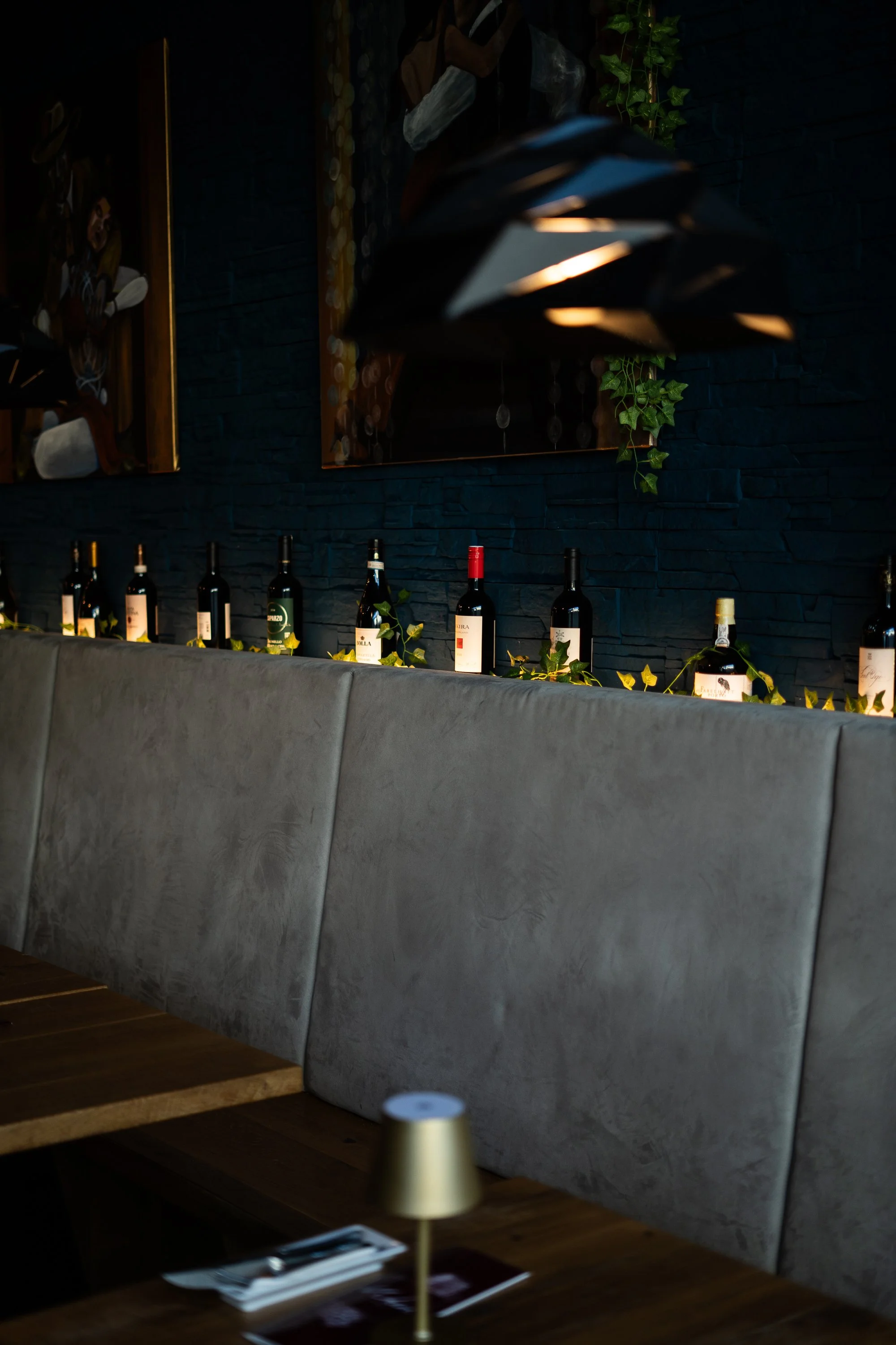 Innenansicht eines Restaurants mit Weinflaschen auf einer erhöhten Ablage gegen eine dunkelblaue Steinwand, dekoriert mit Lichterketten und grünen Kletterpflanzen. Im Vordergrund ein Tisch mit Besteck, einem kleinen goldfarbenen Lampenglas und einer 