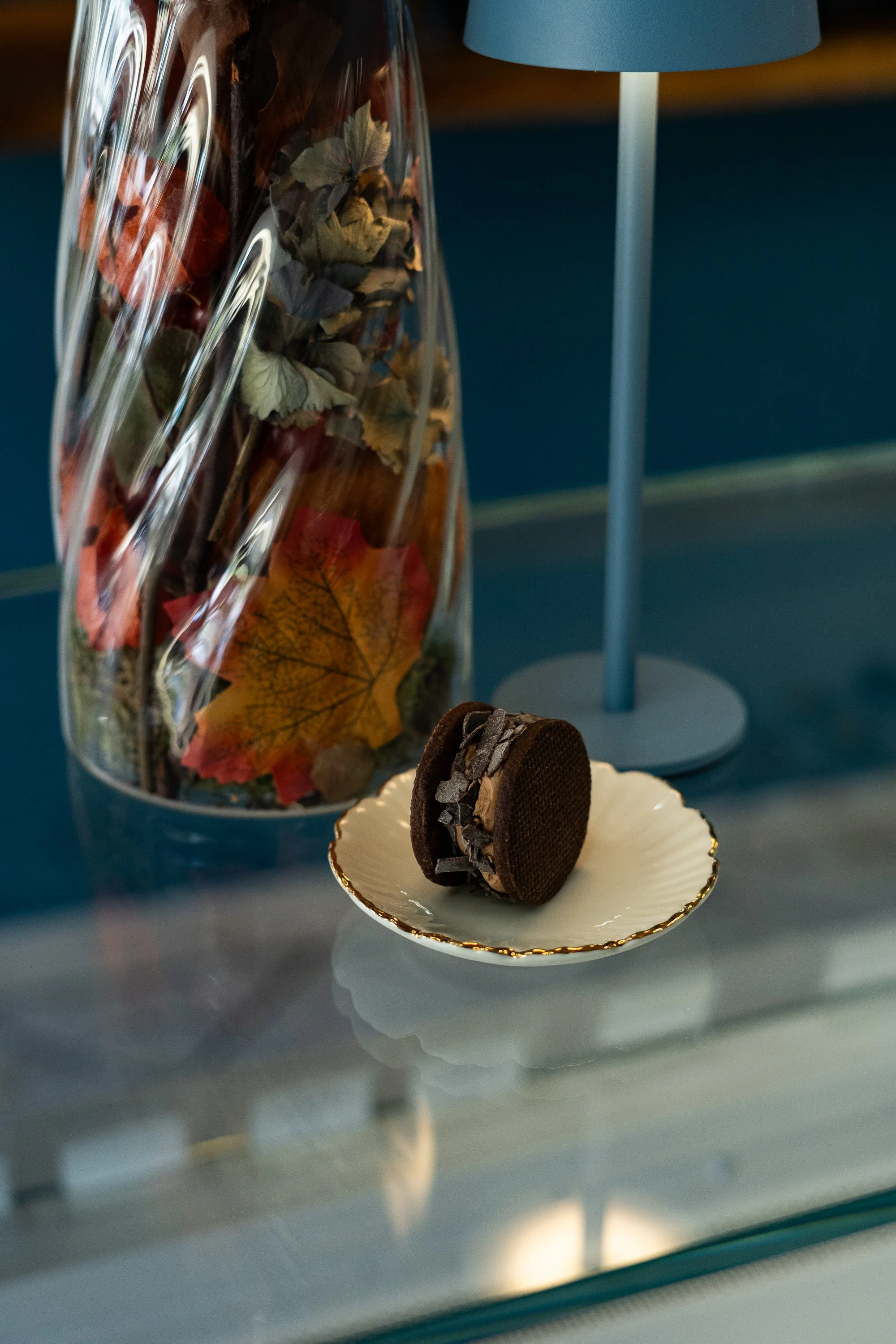 Dunkle Schokoladentorte auf einem weißen Teller mit goldene Kante, im Hintergrund eine Vase mit Herbstblättern und ein blauer Tischlampenschirm.